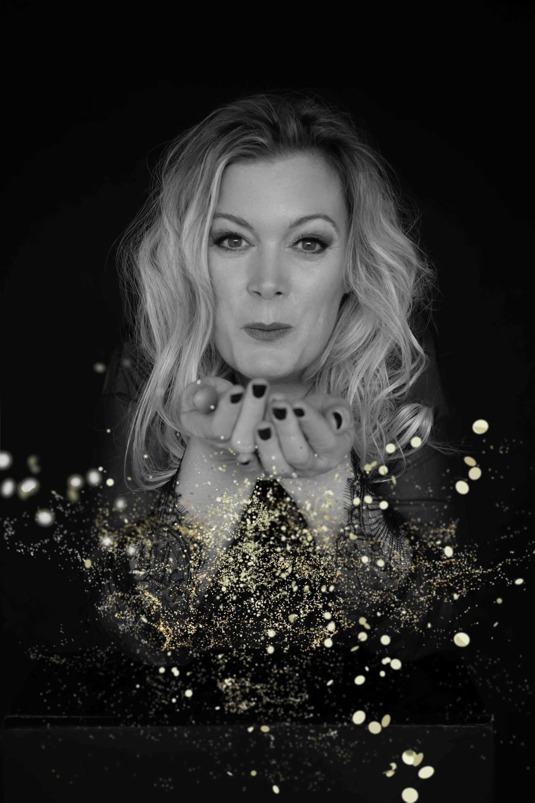 Black and white photo of a woman with wavy hair blowing halo-like golden glitter or light particles from her cupped hands towards the camera against a dark background.