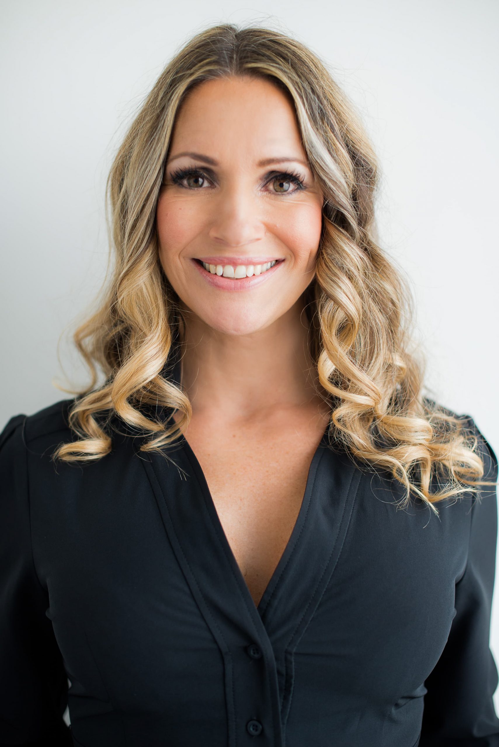 Smiling woman with long blonde curly hair wearing a black buttoned shirt, against a plain white background.