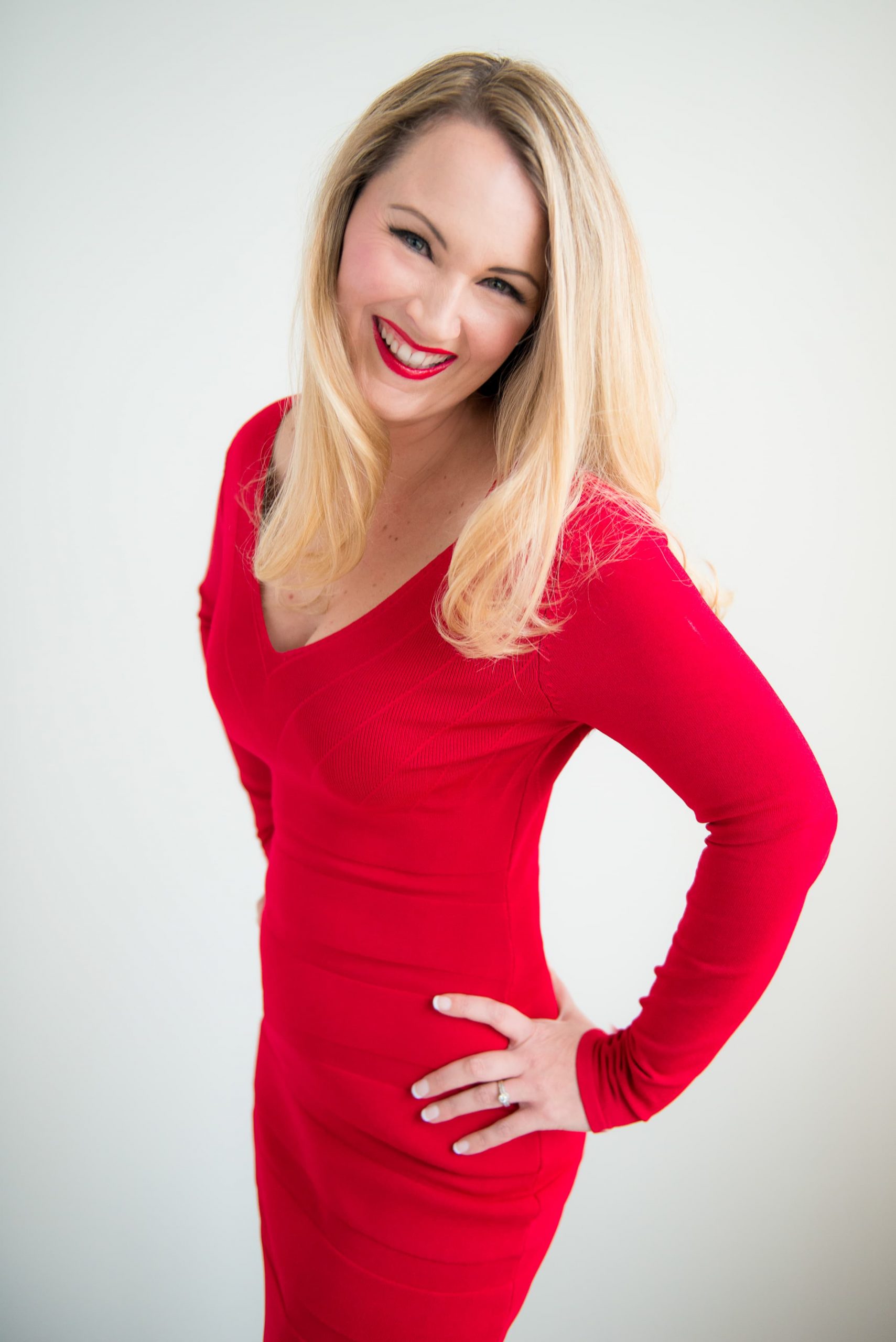 Smiling blonde woman wearing a red long-sleeve dress with one hand on her hip against a plain light background.