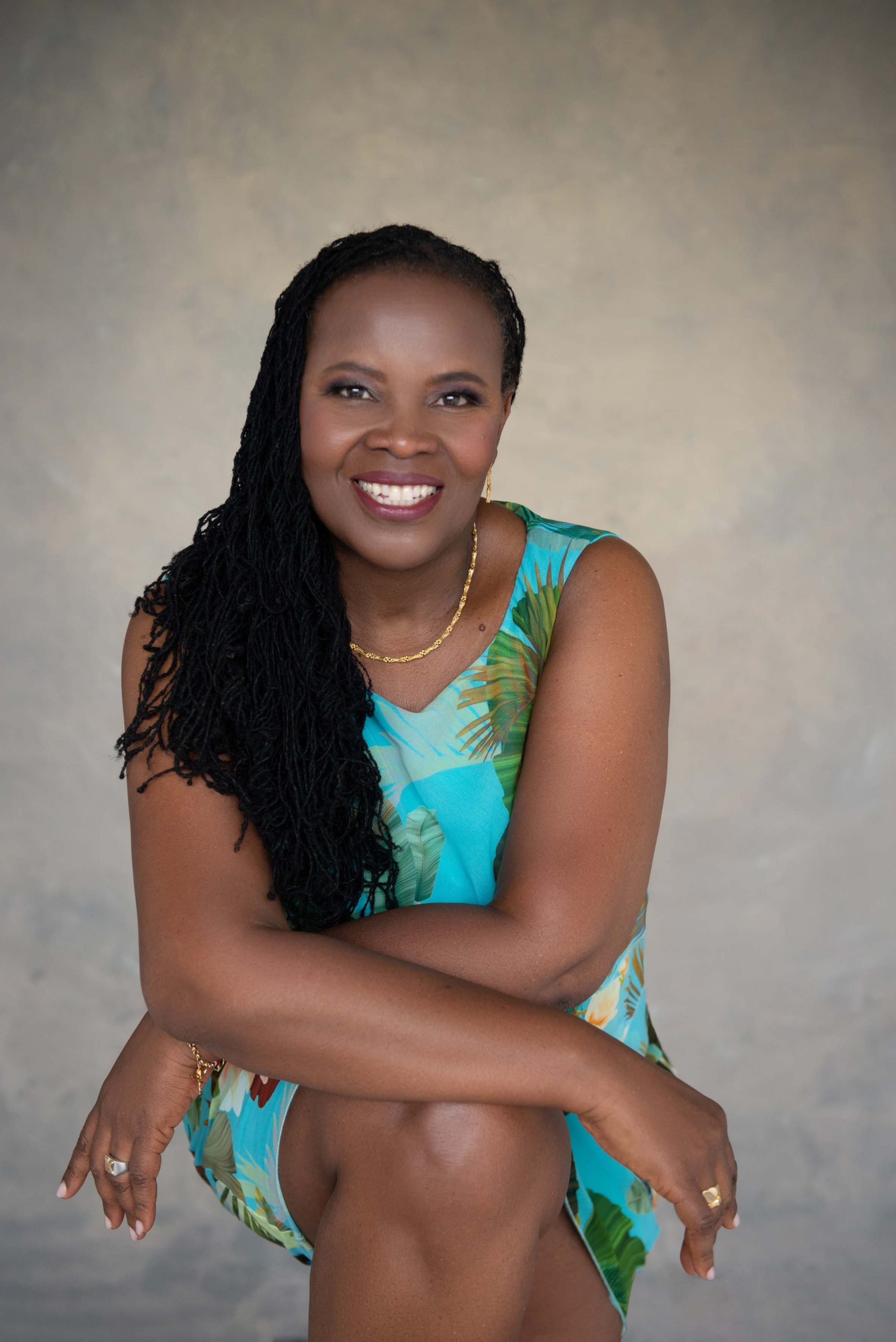Smiling woman with long braided hair wearing a blue sleeveless dress with green leaf patterns, seated with her arms crossed over one knee against a neutral background.