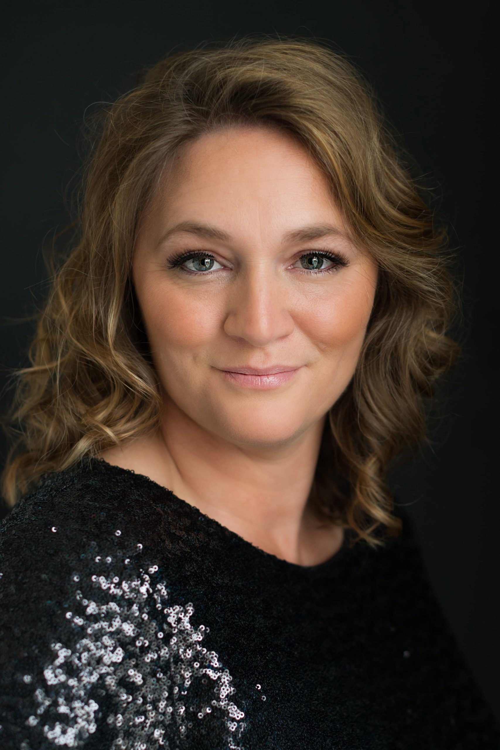 Close-up portrait of a woman with light brown curly hair, green eyes, and a subtle smile, wearing a black sequin top against a dark background.