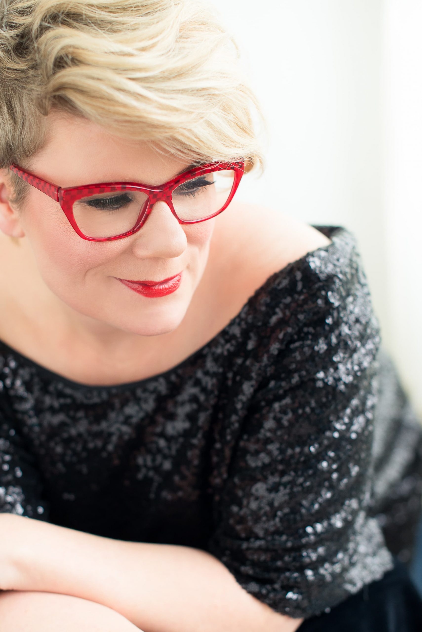 Close-up of a smiling woman with short blonde hair wearing red glasses and red lipstick, dressed in a black sequined off-the-shoulder top.