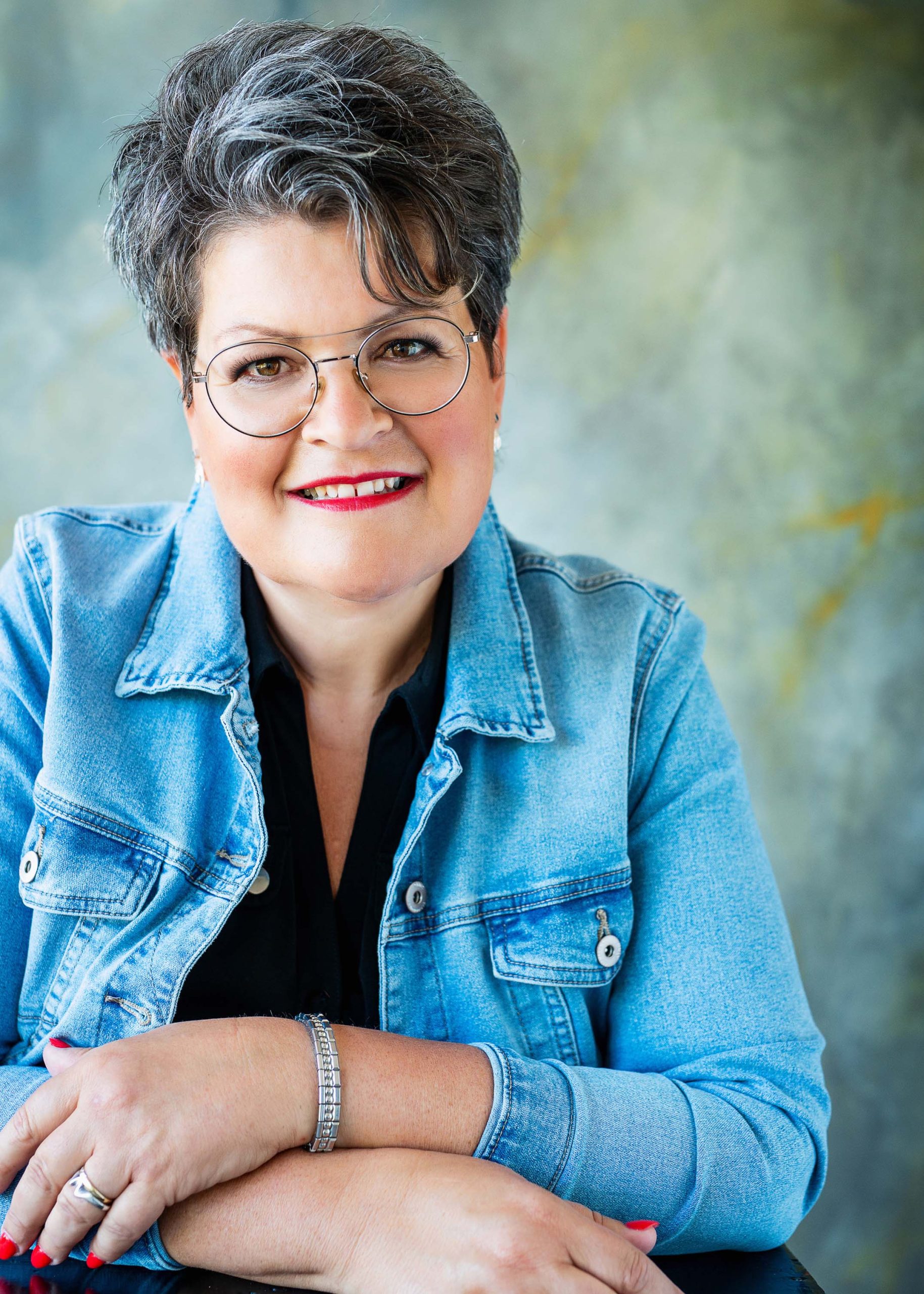 Smiling middle-aged woman with short salt-and-pepper hair, wearing glasses, a light blue denim jacket, and a black shirt, resting her crossed arms on a surface.