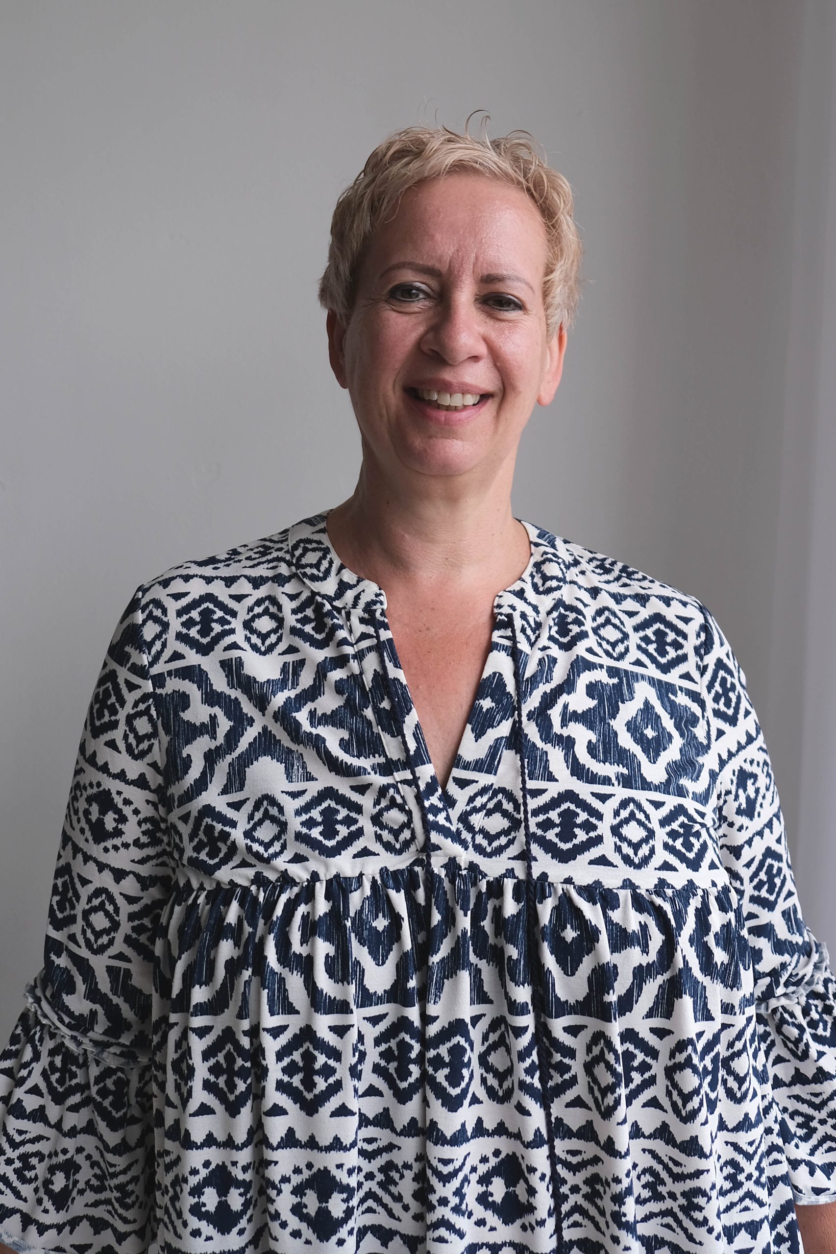 Smiling middle-aged person with short blonde hair wearing a white and navy blue patterned long-sleeve blouse standing against a plain light gray background.