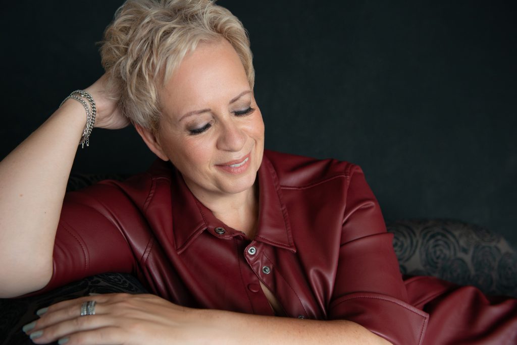 Woman with short blonde hair wearing a burgundy leather jacket, resting her head on her left hand with eyes closed and smiling gently.