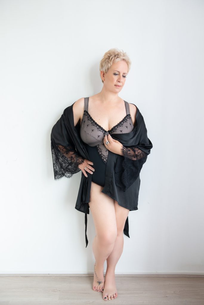 A woman with short blonde hair stands barefoot against a white wall wearing black lace lingerie and a matching black robe with lace sleeves, looking downward with her left hand resting on her hip and her right hand holding the robe closed.