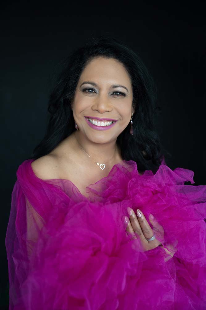 Smiling woman with long black hair wearing a bright pink tulle garment and a silver necklace with two interlocked hearts, standing against a black background.