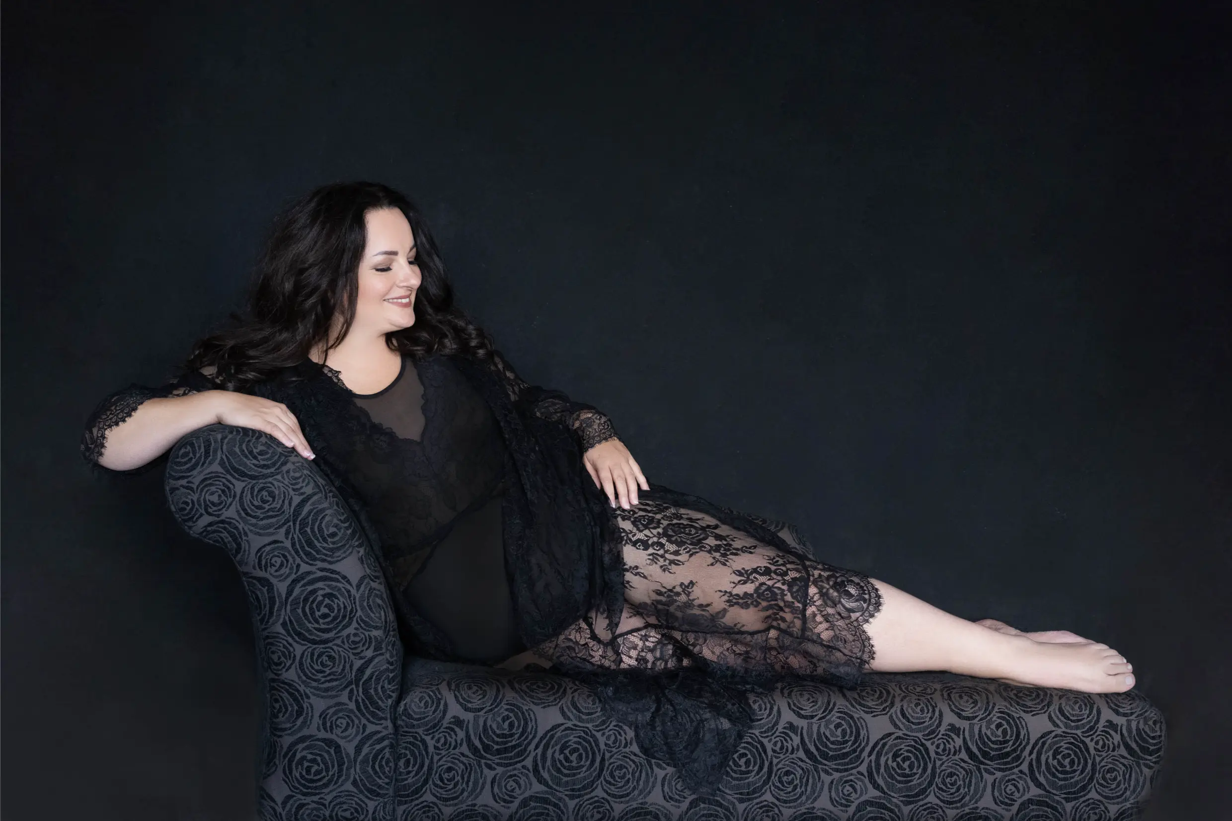 A smiling woman with long dark hair reclines on a chaise lounge covered in a black rose-patterned fabric. She wears a sheer black lace dress and is barefoot against a solid black background.