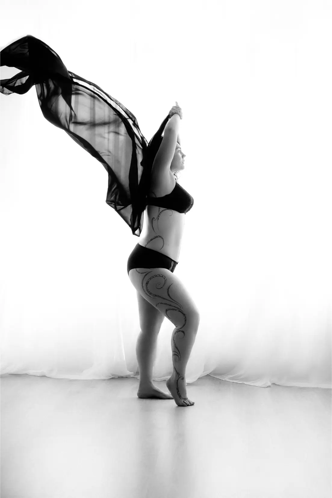 Black and white photo of a woman standing on one leg, wearing black lingerie and holding a flowing sheer fabric behind her, with decorative tattoos on her side and leg.