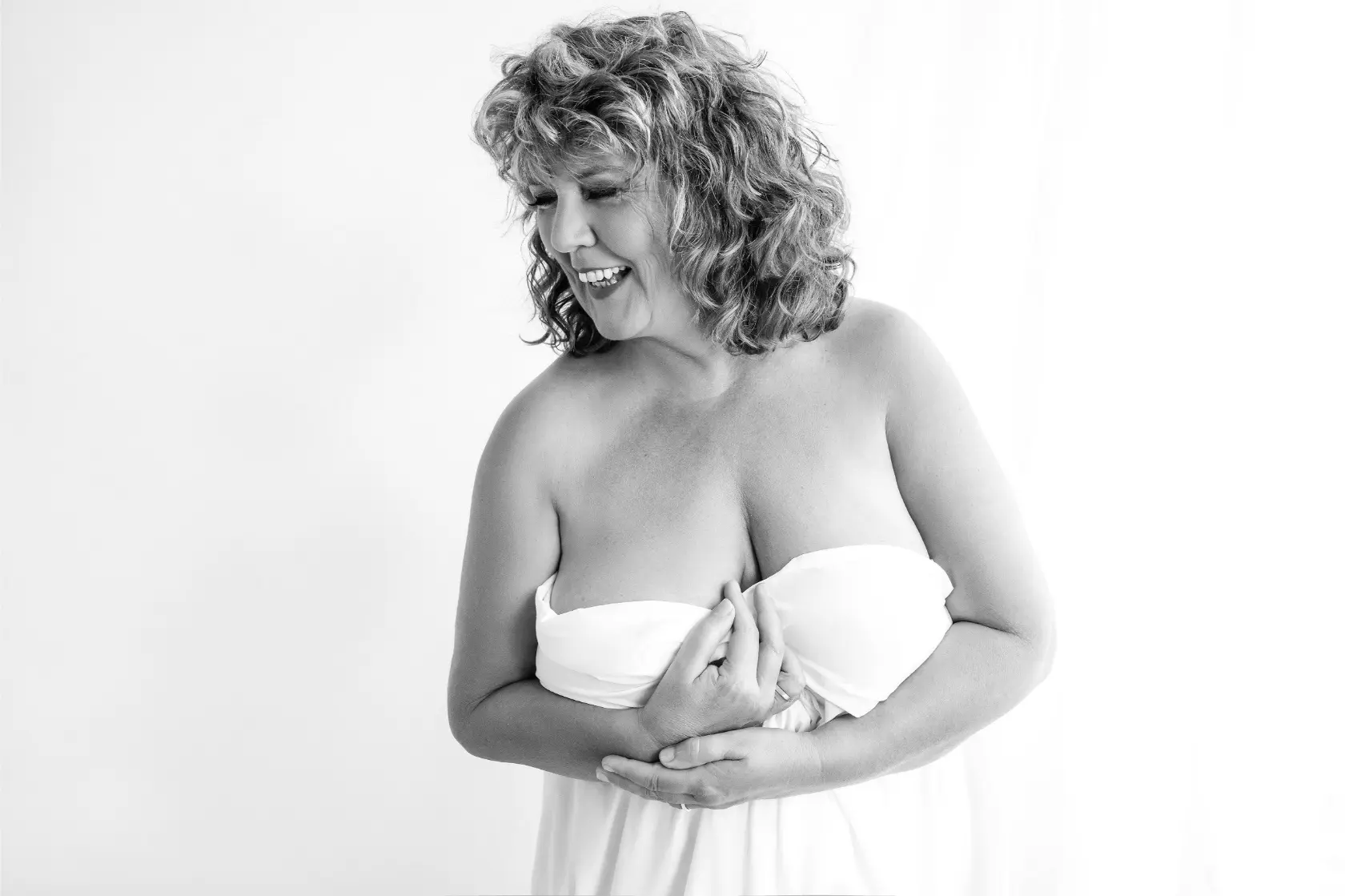 Black and white photo of a smiling middle-aged woman with curly hair, wearing a strapless white dress, looking down to her left while holding the front of the dress with both hands.