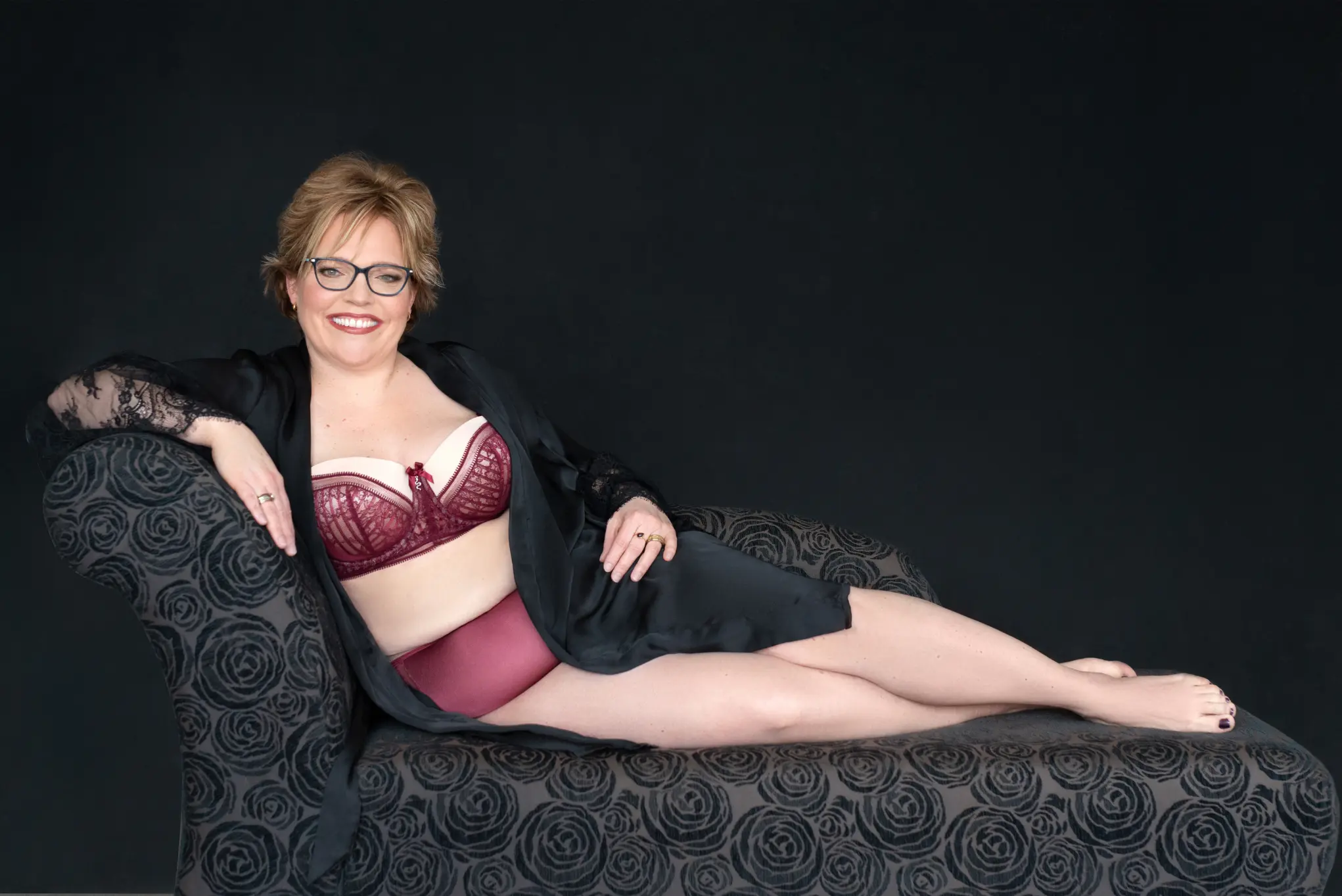 Smiling woman with short blonde hair and glasses reclining on a black floral-patterned chaise lounge, wearing a burgundy lace bra, matching high-waisted underwear, and a black sheer robe with lace sleeves, against a plain dark background.