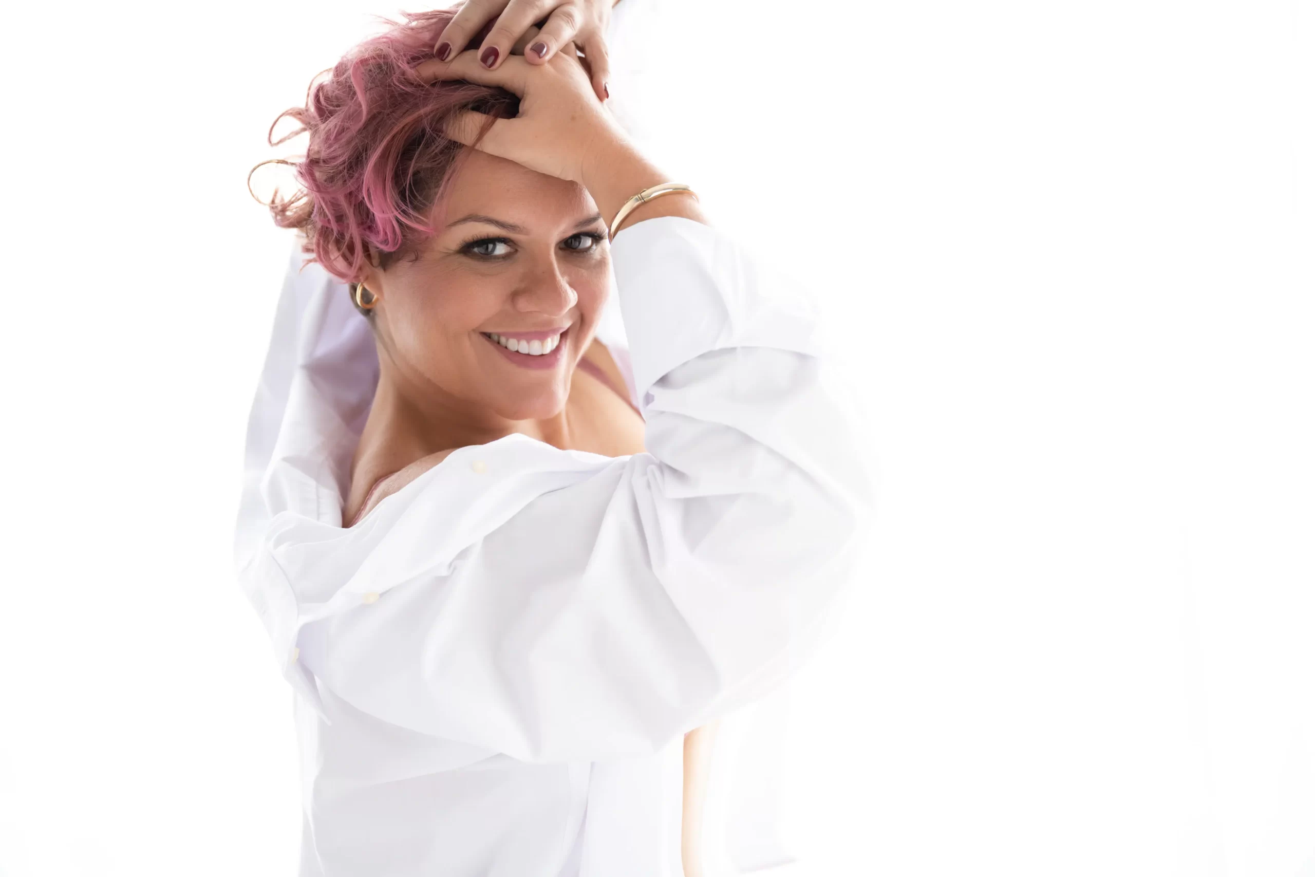 Smiling woman with short curly pink hair wearing a white shirt, posed with her hands on her head and looking at the camera.