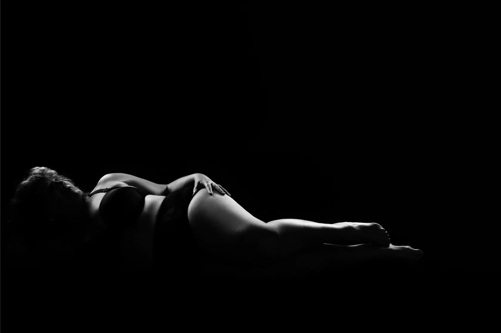 Black and white silhouette of a woman lying on her side in lingerie against a dark background, softly lit to highlight curves.