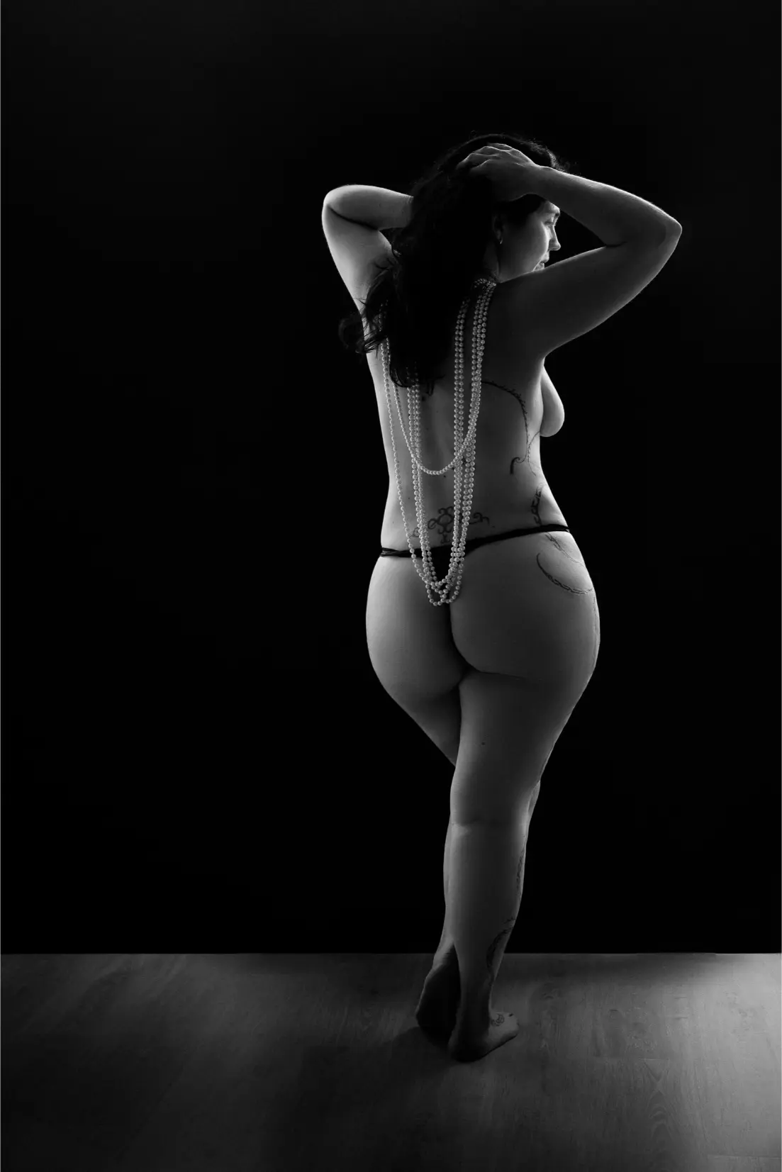 Black and white photo of a nude woman with curvy body and long dark hair standing on wooden floor against a black background, seen from behind, wearing long strands of pearls draped down her back and holding her hair with both hands, showing tattoos on her side and leg.