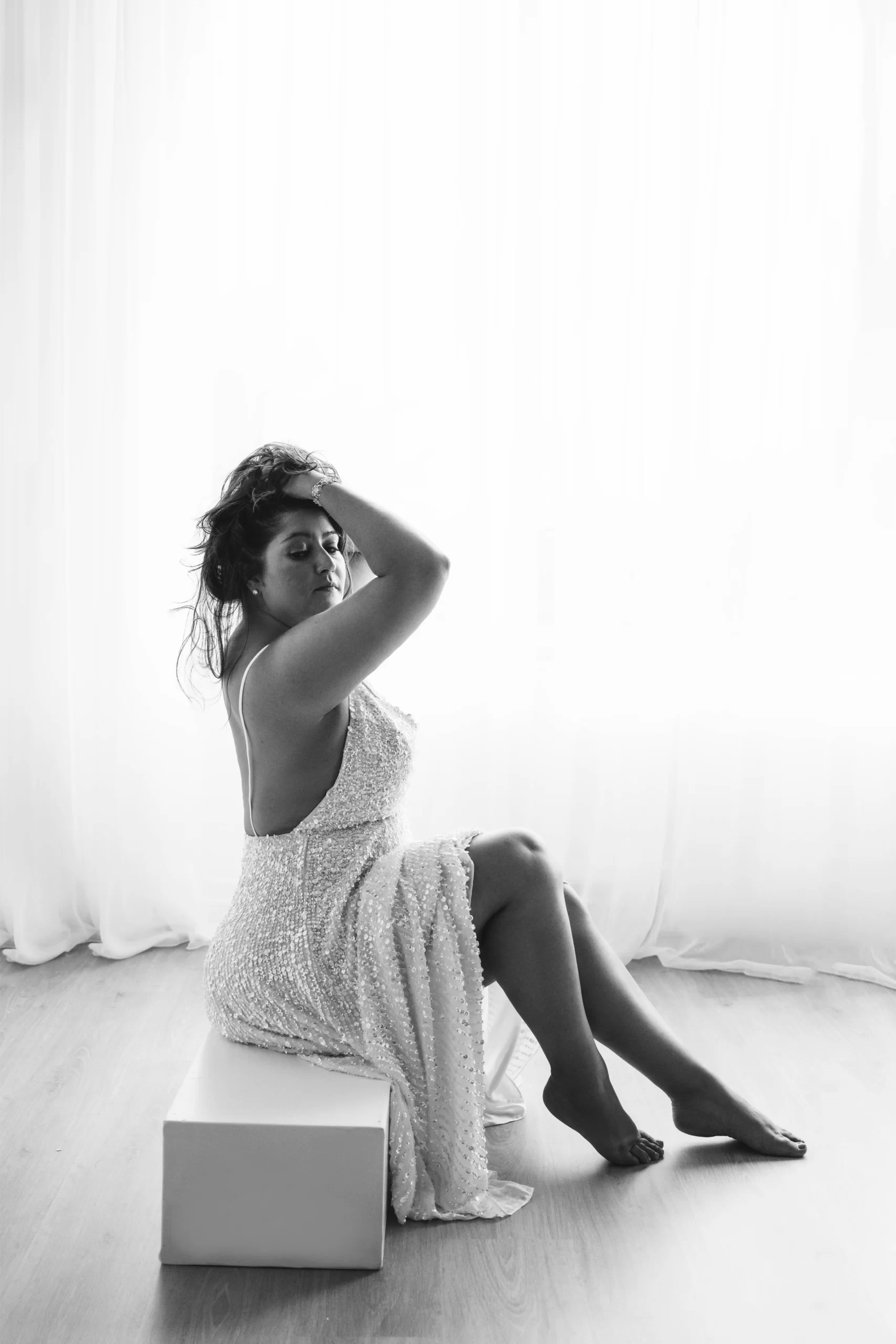 Black-and-white photo of a woman in a sleeveless, textured dress sitting on a white cube, with one arm raised to touch her hair and legs extended on a wooden floor.