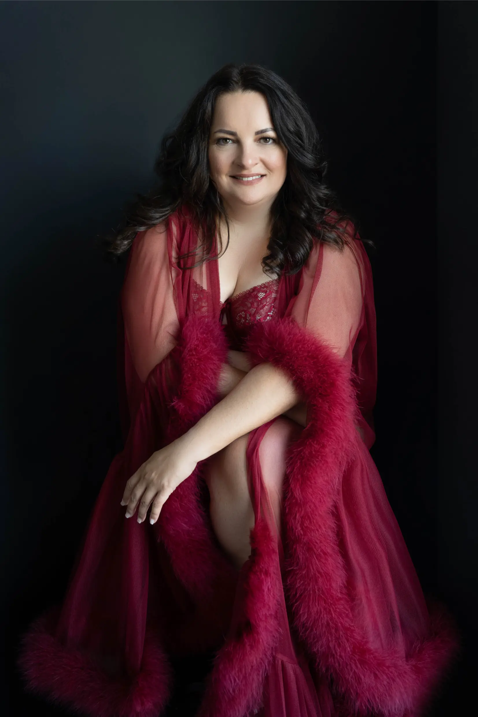Smiling woman with long dark hair wearing a sheer red robe with fluffy trim and matching lace lingerie, sitting with arms crossed over her bent knee against a dark background.