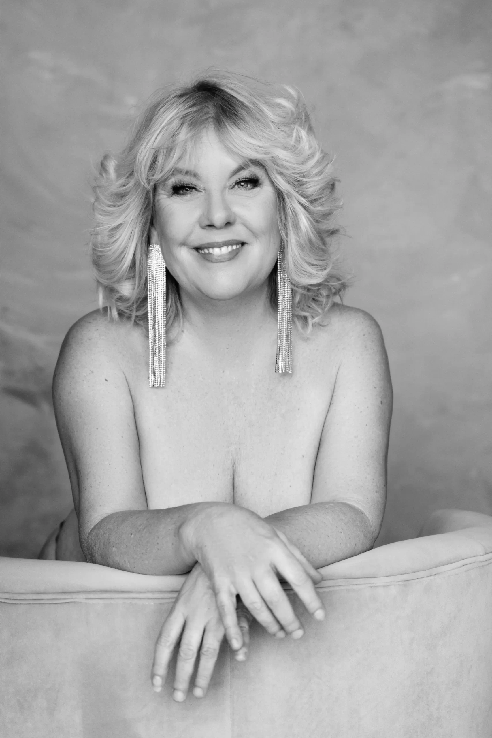 Black and white portrait of a smiling middle-aged woman with blonde hair styled in soft waves, wearing long, dangling earrings, leaning on a cushioned surface with her arms crossed.
