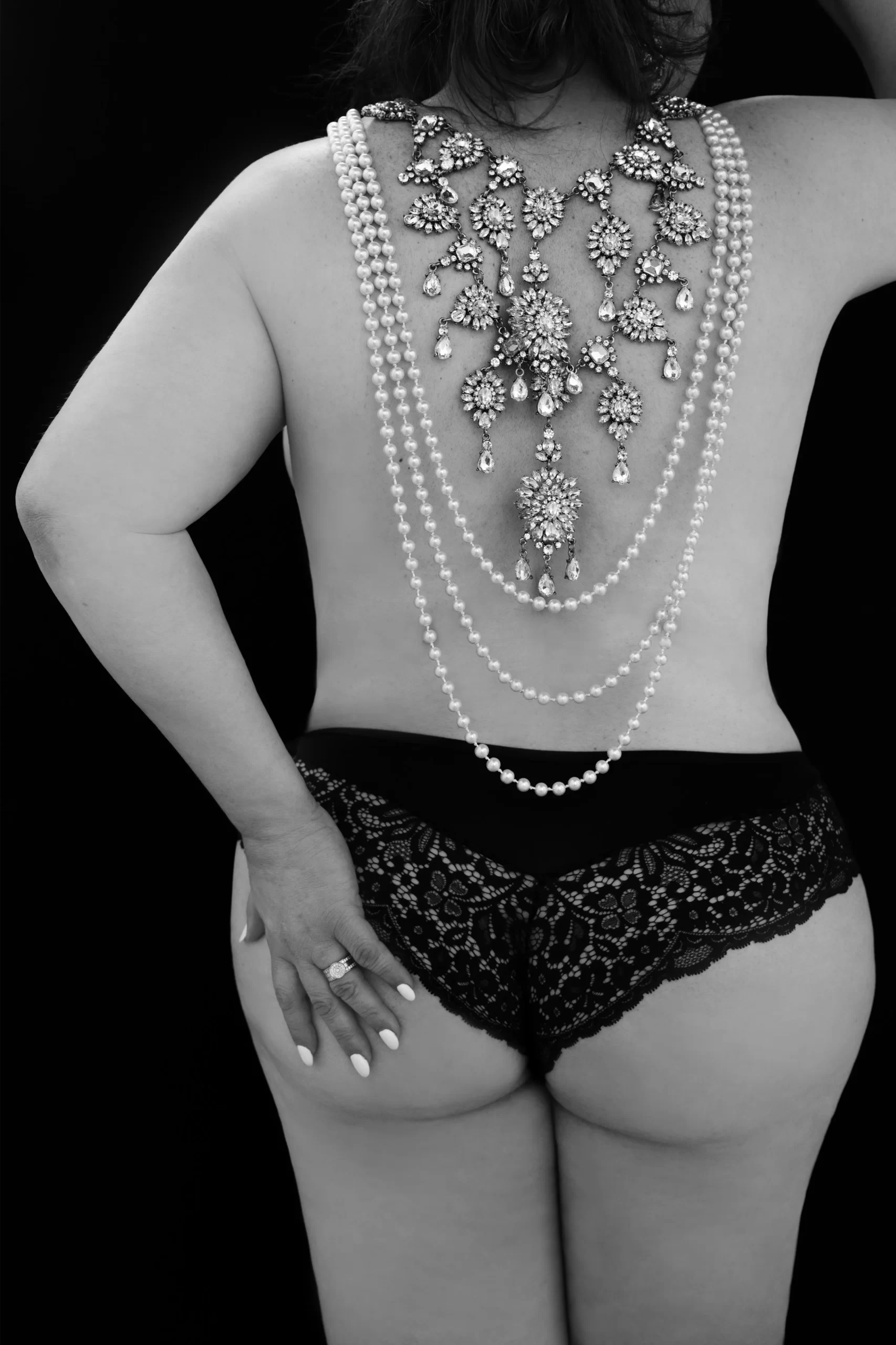 Black and white photo showing a woman from the back wearing black lace underwear and an elaborate pearl and jewel necklace draped down her bare back, with her left hand resting on her hip displaying manicured nails and a ring.