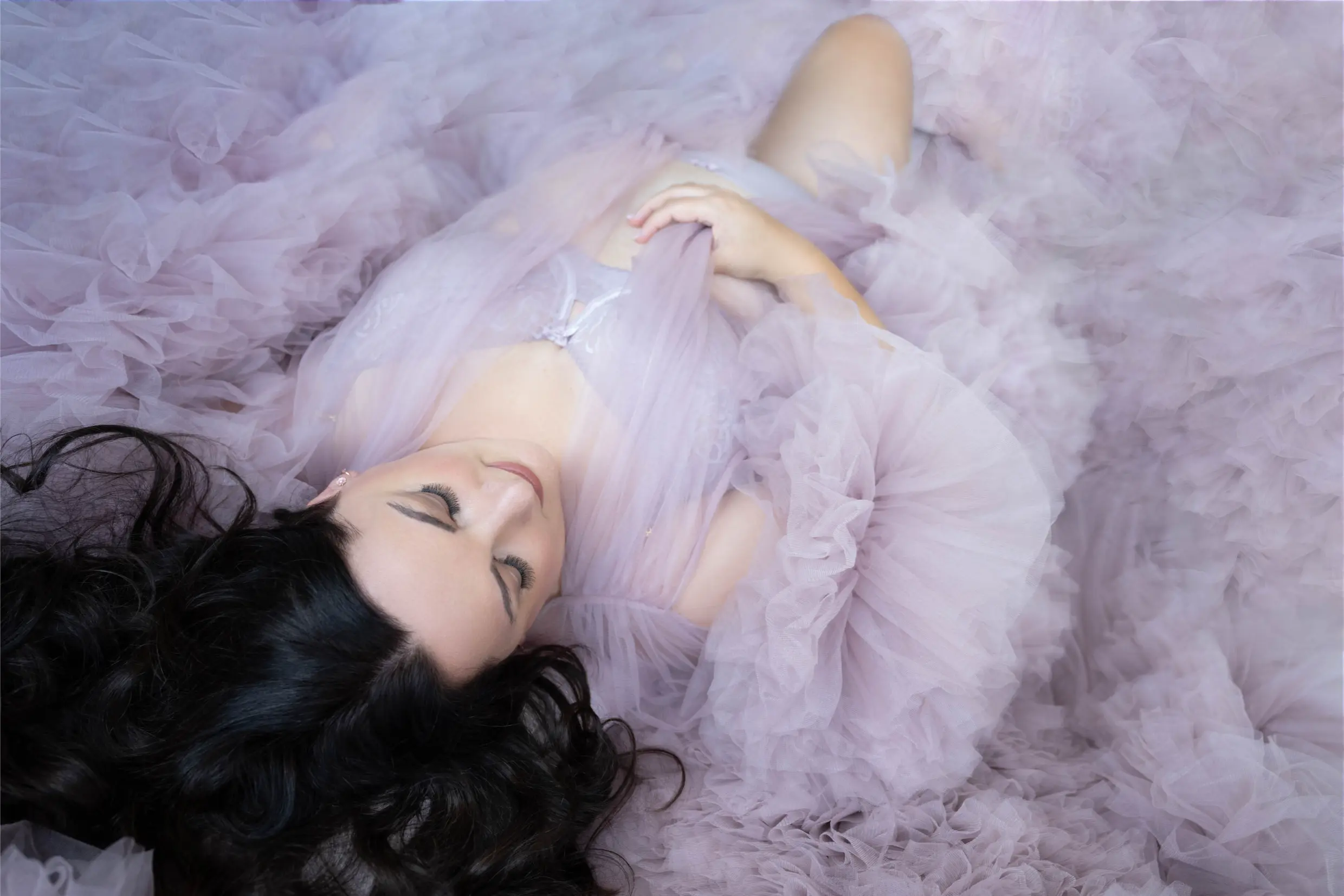 Woman with long dark hair lying down with eyes closed, surrounded by layers of soft, light pink tulle fabric.