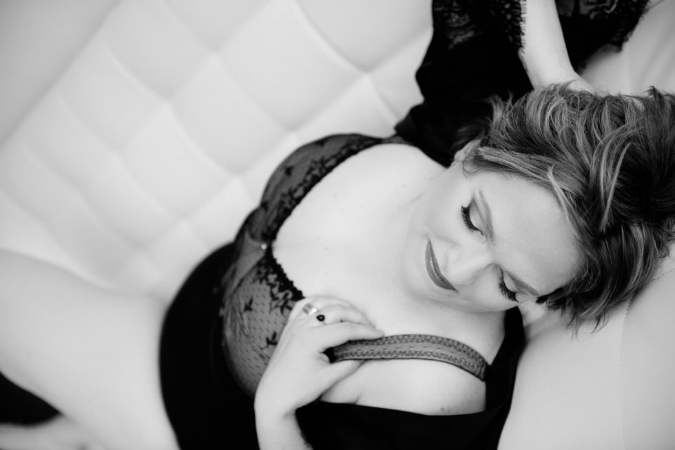 Black and white image of a woman reclining on a quilted sofa, wearing a lace lingerie outfit with one strap off her shoulder, looking down with a gentle smile.