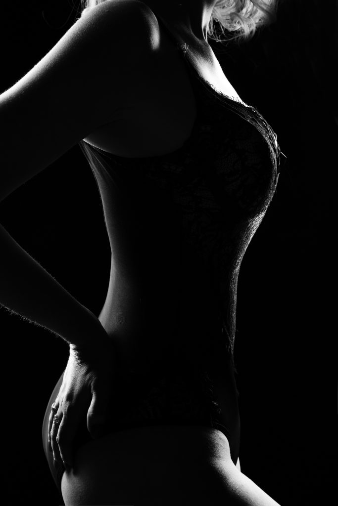 Black and white side-profile silhouette of a woman wearing lace lingerie with her hand resting on her hip, highlighting her curves against a dark background.