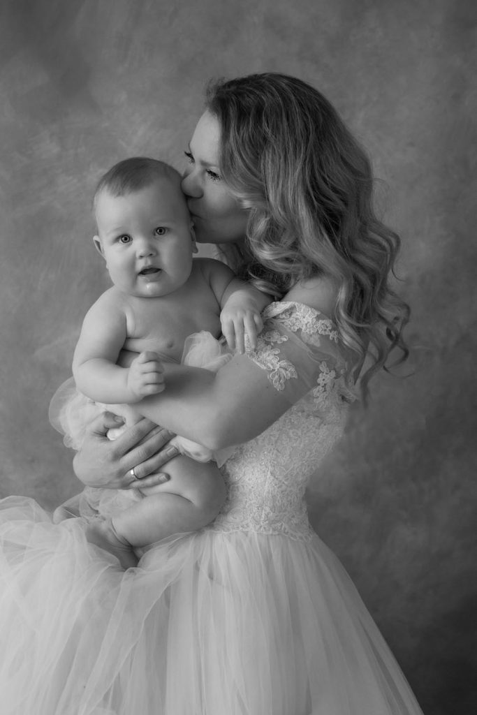 Black and white portrait of a woman in an off-shoulder lace dress holding and kissing a baby wearing a tulle skirt, against a soft textured backdrop.