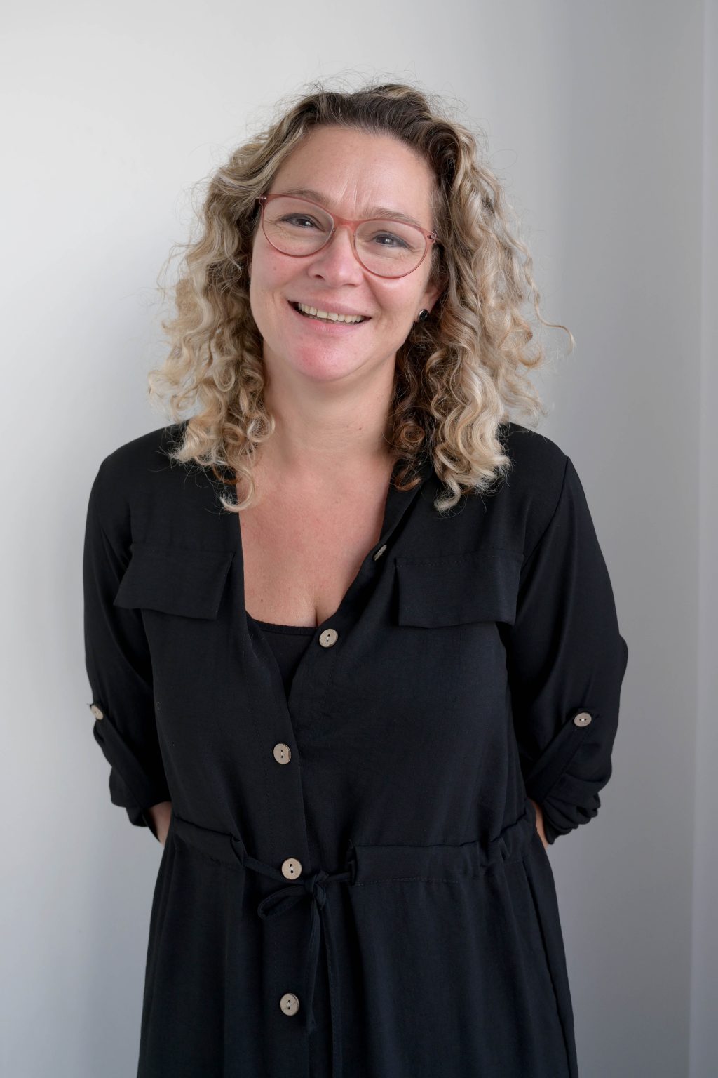 Smiling woman with curly blonde hair wearing red glasses and a black buttoned dress standing against a plain light gray background.