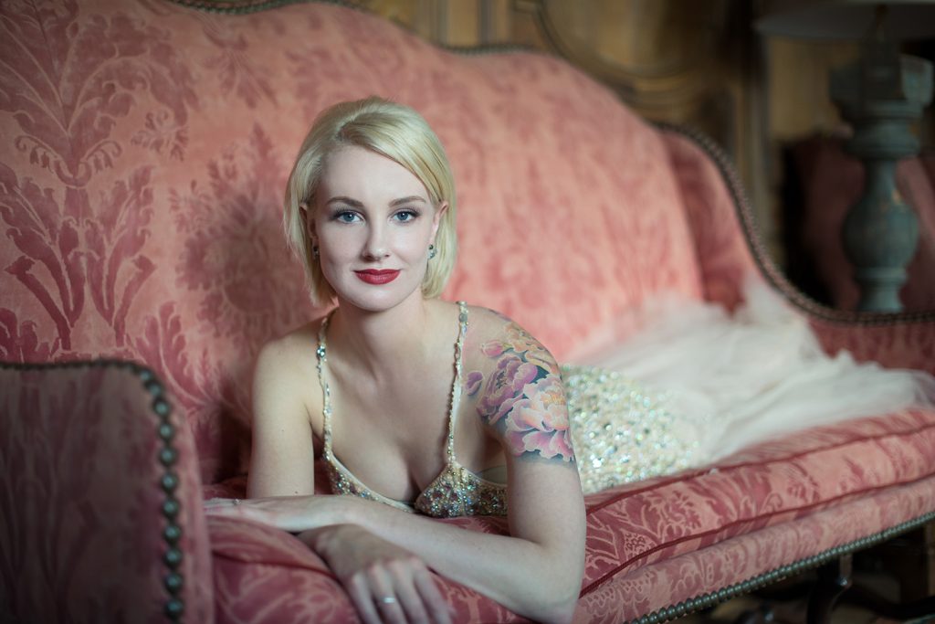 Blonde woman with a colorful floral tattoo on her right shoulder, wearing a sparkling dress with thin straps, reclining on a pink patterned vintage sofa and smiling at the camera.