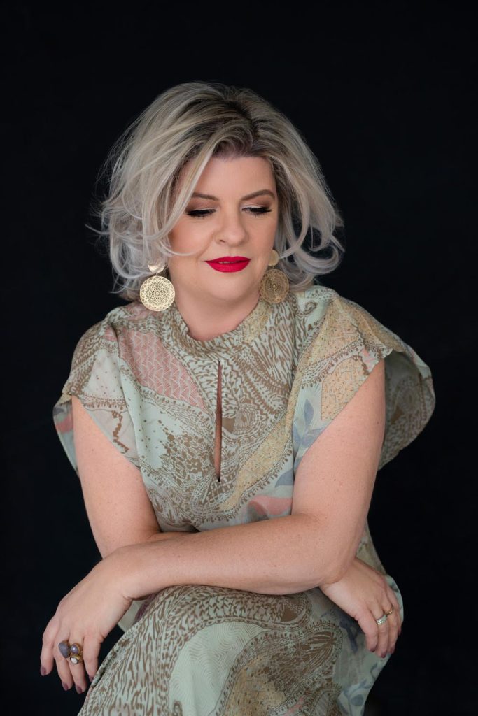 Woman with short wavy blonde hair wearing a patterned pastel dress, large round gold earrings, and bright red lipstick, sitting with her arms crossed and looking down against a black background.