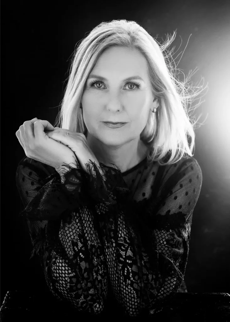 Black and white portrait of a mature woman with light hair and clear eyes wearing a patterned lace top, resting her chin on her clasped hands, with light softly illuminating her hair from behind.
