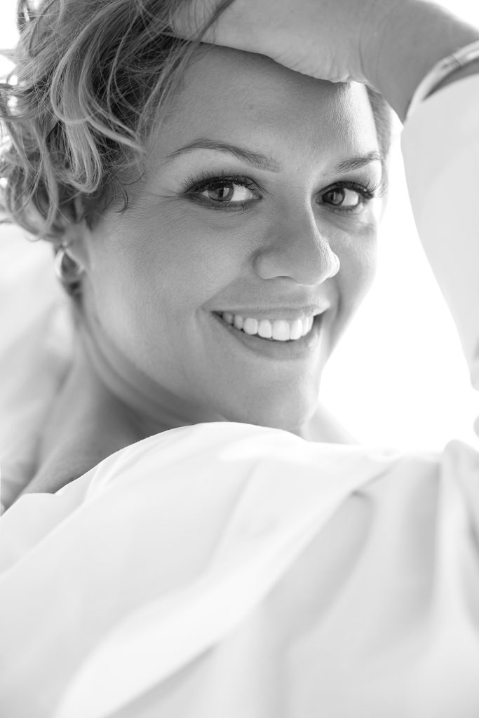 Black and white close-up of a smiling woman with short wavy hair resting her arm on her forehead, wearing a light-colored top.