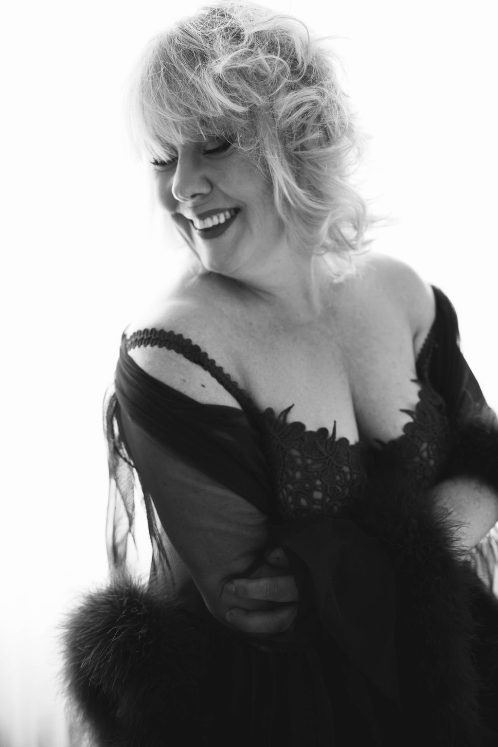 Black and white photo of a smiling woman with short curly hair wearing a lacy, off-the-shoulder dress and sheer robe with feathered cuffs, looking down and to her left while hugging herself.