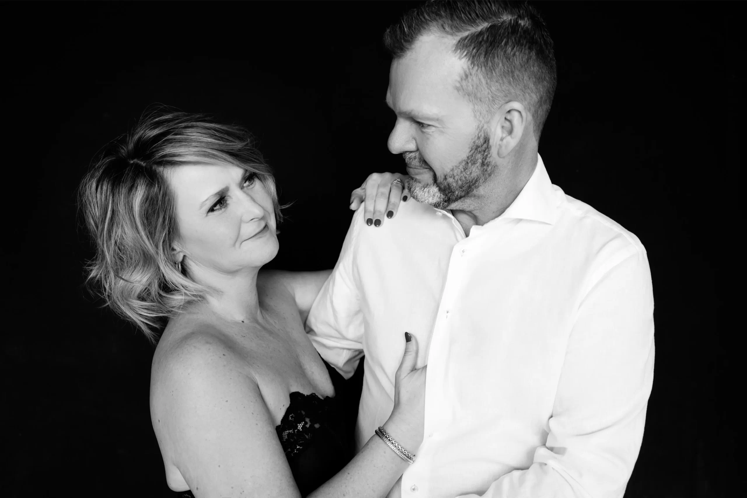 Black and white photo of a woman in a strapless lace top looking lovingly at a man in a white shirt who returns her gaze with a gentle smile, her hand resting on his shoulder and arm.