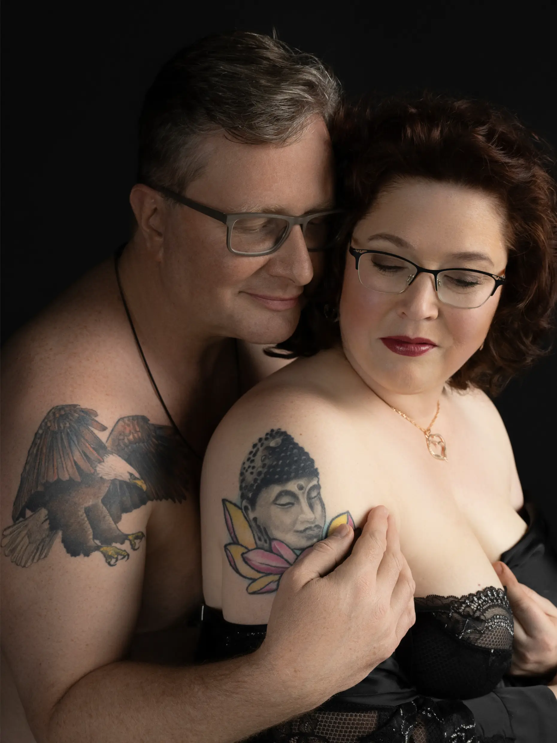 A couple with glasses posing intimately against a black background, the man embracing the woman from behind while both show prominent tattoos on their upper arms; the man has an eagle tattoo and the woman a Buddha with lotus flowers tattoo.