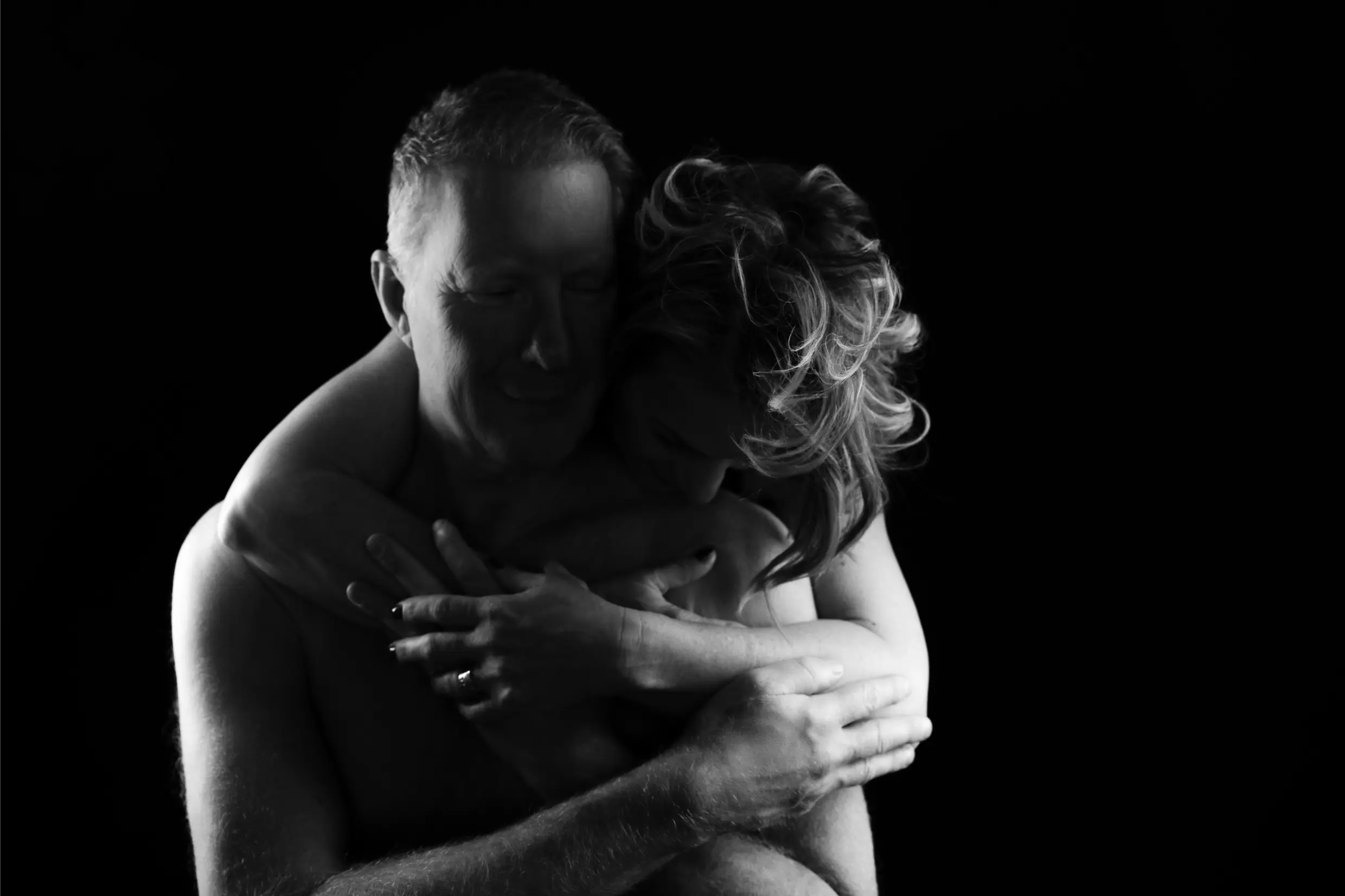 Black and white photo of a shirtless man and woman embracing against a black background, the woman wrapping her arms around the man from behind with her head resting on his shoulder.