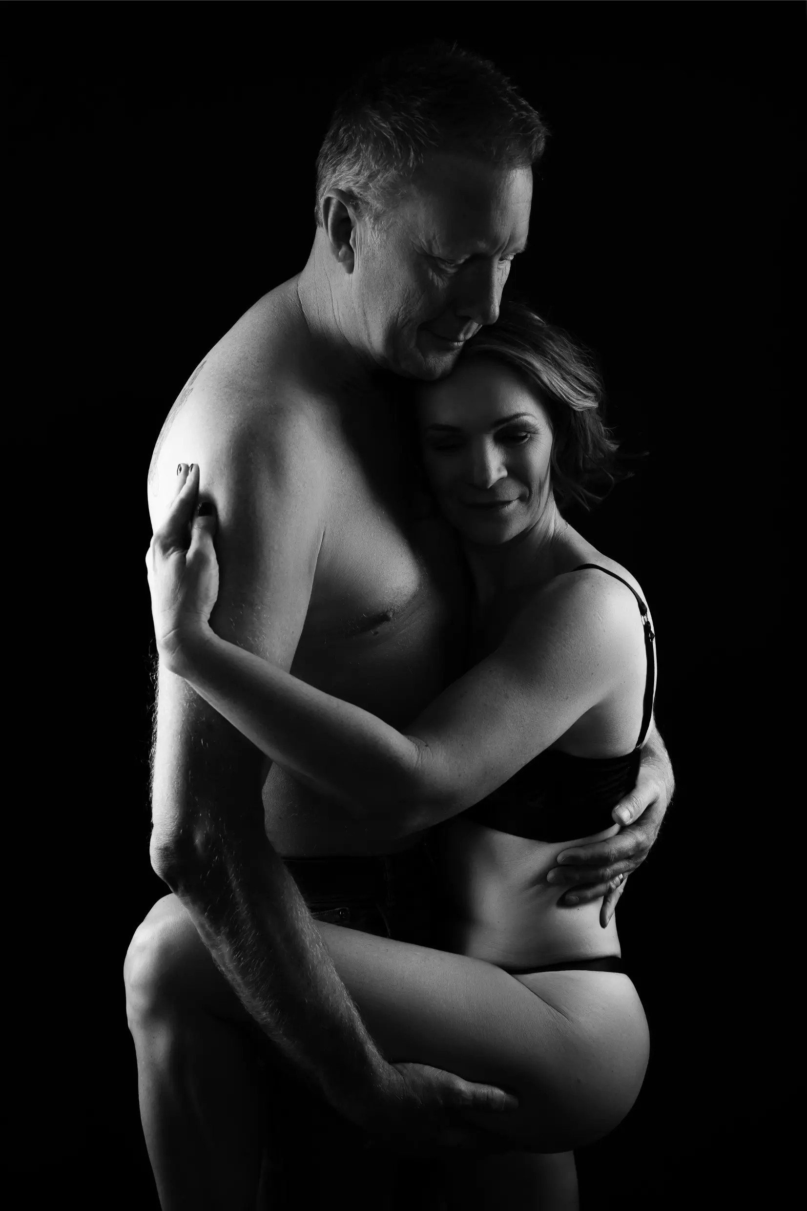 Black and white intimate portrait of a shirtless man embracing a woman in black lingerie against a black background, both with eyes closed and smiling gently.