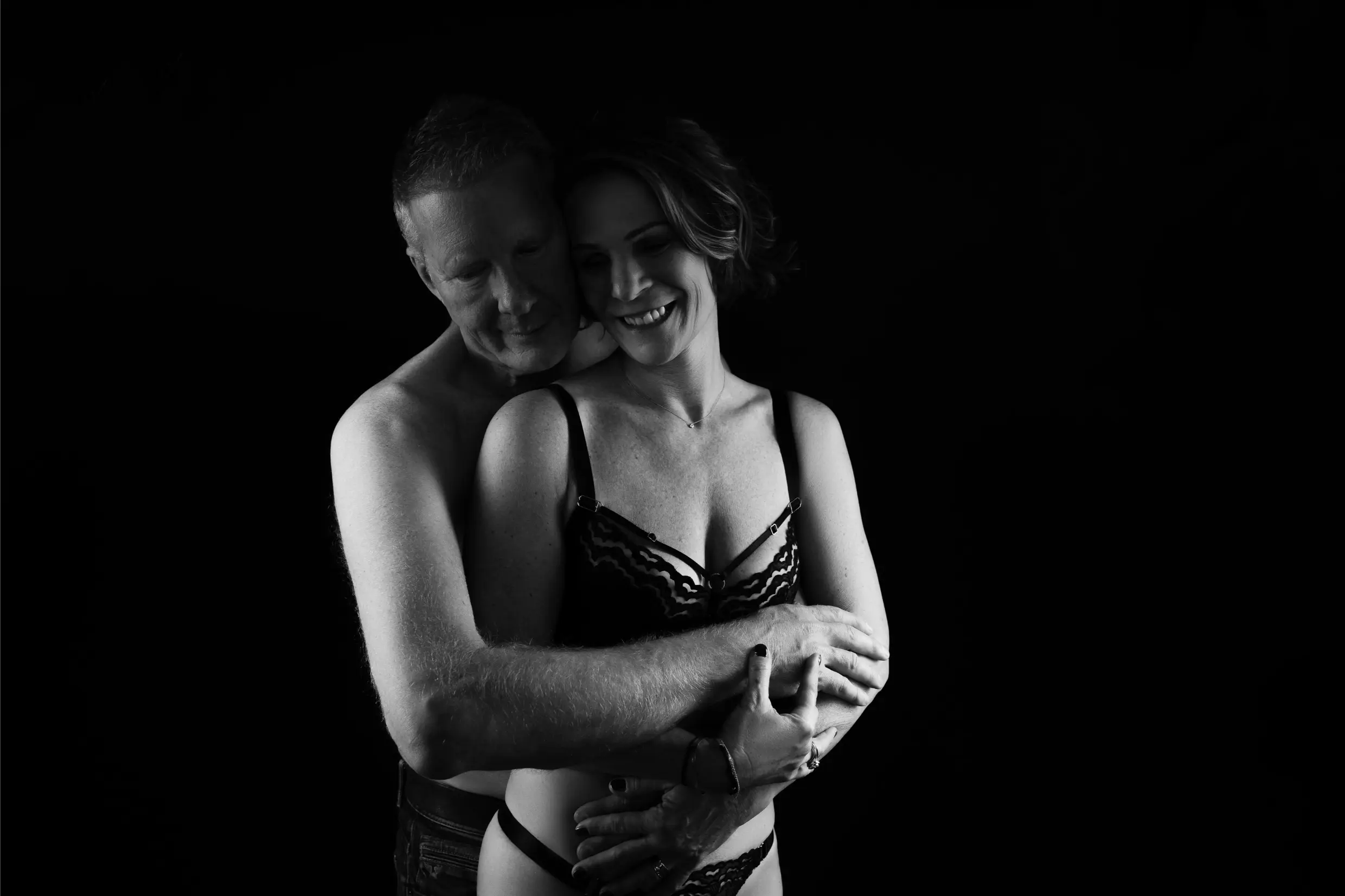 Black and white photo of a shirtless man embracing a smiling woman in lace lingerie from behind against a dark background.