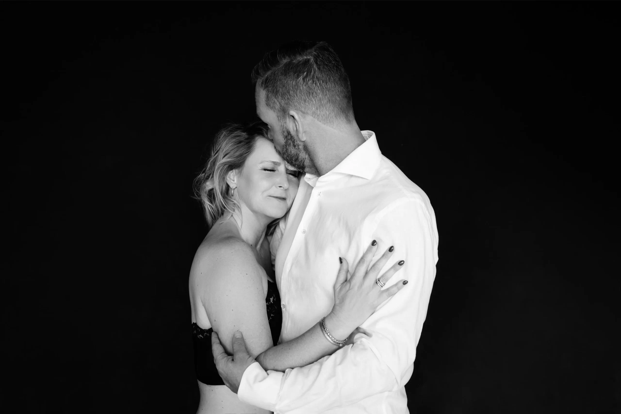 Black and white photo of a couple embracing tenderly against a black background; the woman with closed eyes wears a strapless top and the man in a white shirt gently kisses her forehead.