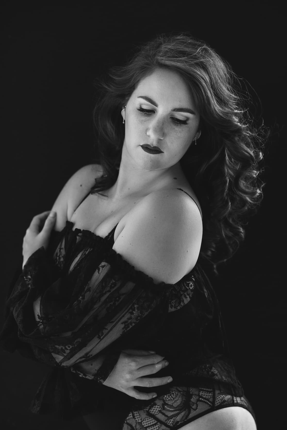 Black and white portrait of a woman with wavy hair wearing a lace off-the-shoulder top and looking down with a pensive expression against a dark background.