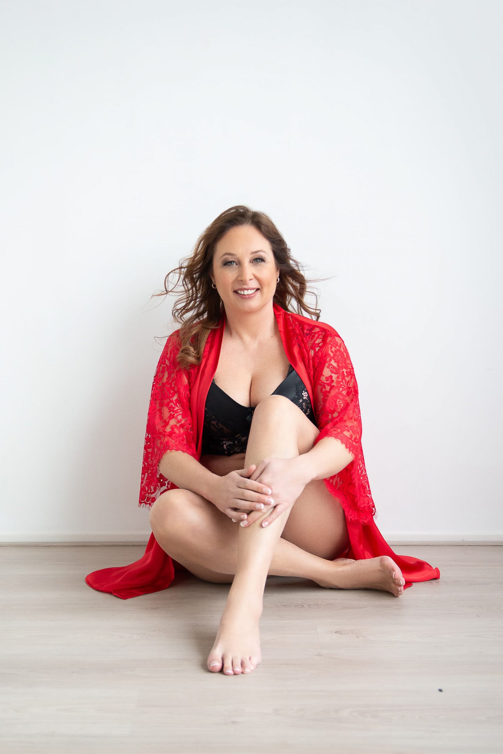 Smiling woman with wavy brown hair sitting barefoot on a light wood floor against a white wall, wearing a black lace bra and a red lace robe.