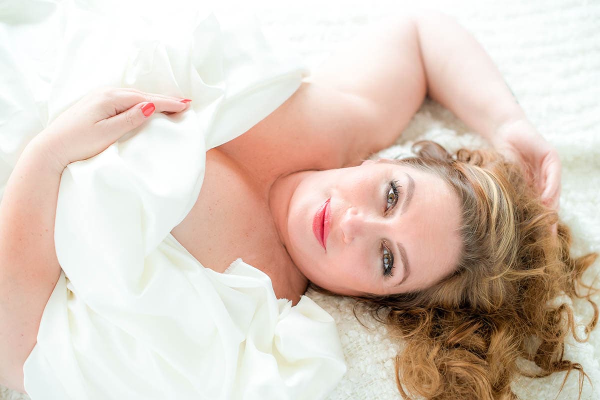 Woman with wavy light brown hair lying on a white textured surface, partially covered by a white sheet, looking up at the camera with a soft smile.