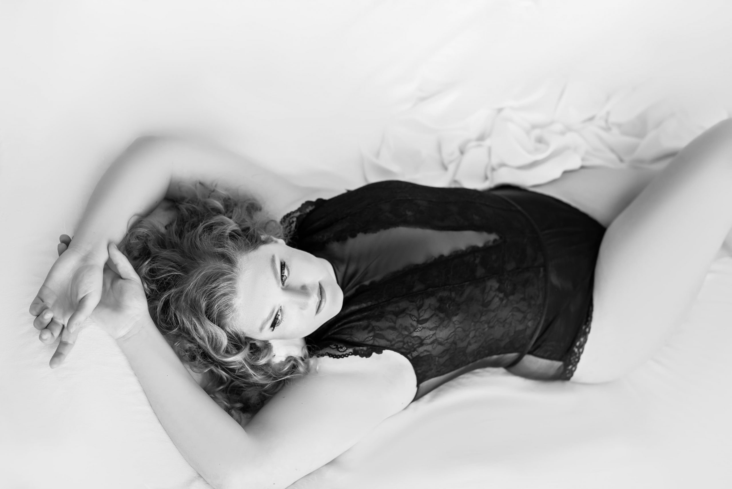 Black and white photograph of a woman with curly hair lying on a bed wearing a black lace bodysuit, with her arms stretched above her head and looking slightly upwards.