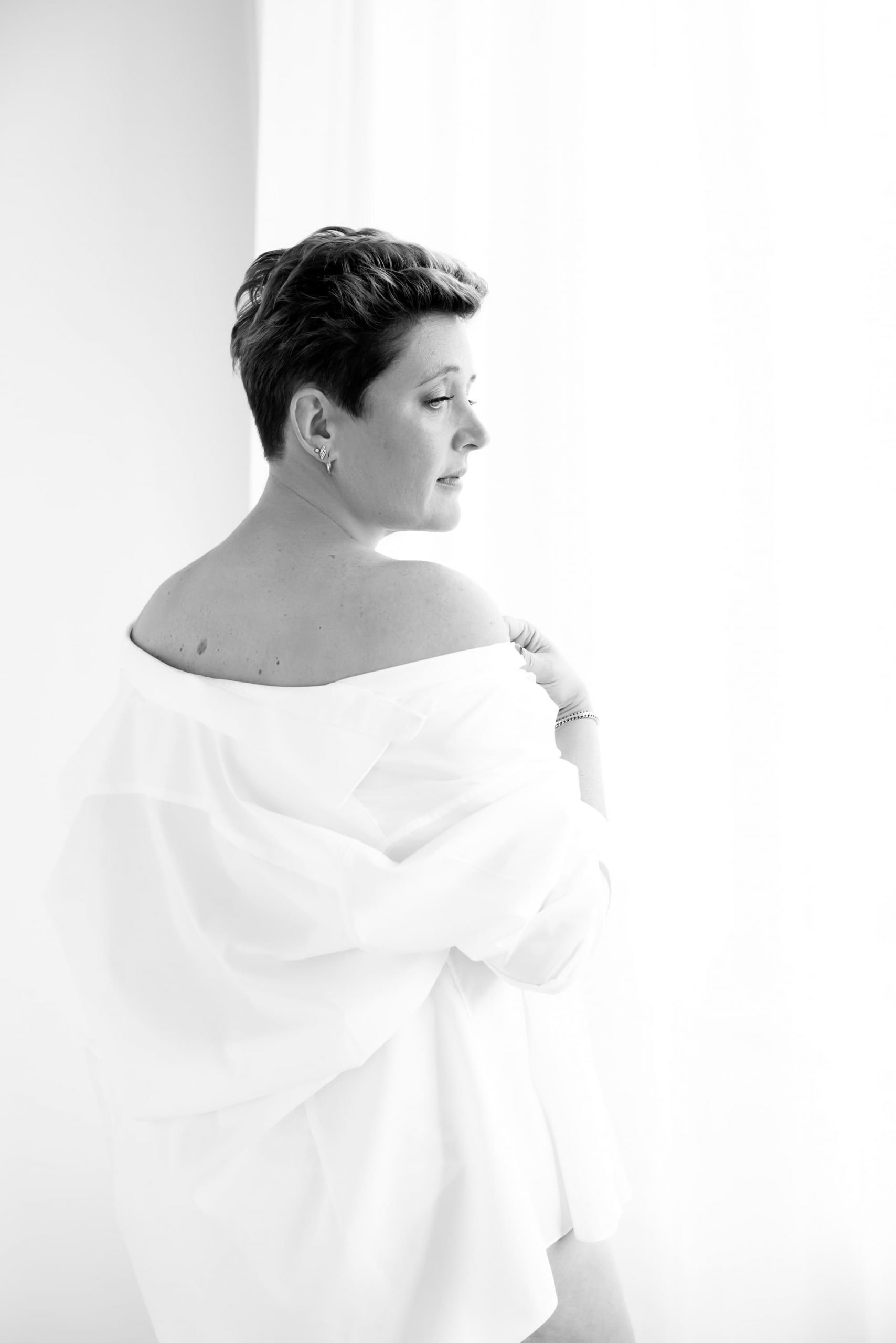 Black and white portrait of a short-haired woman viewed from behind, wearing an off-shoulder white shirt, looking towards a bright window.
