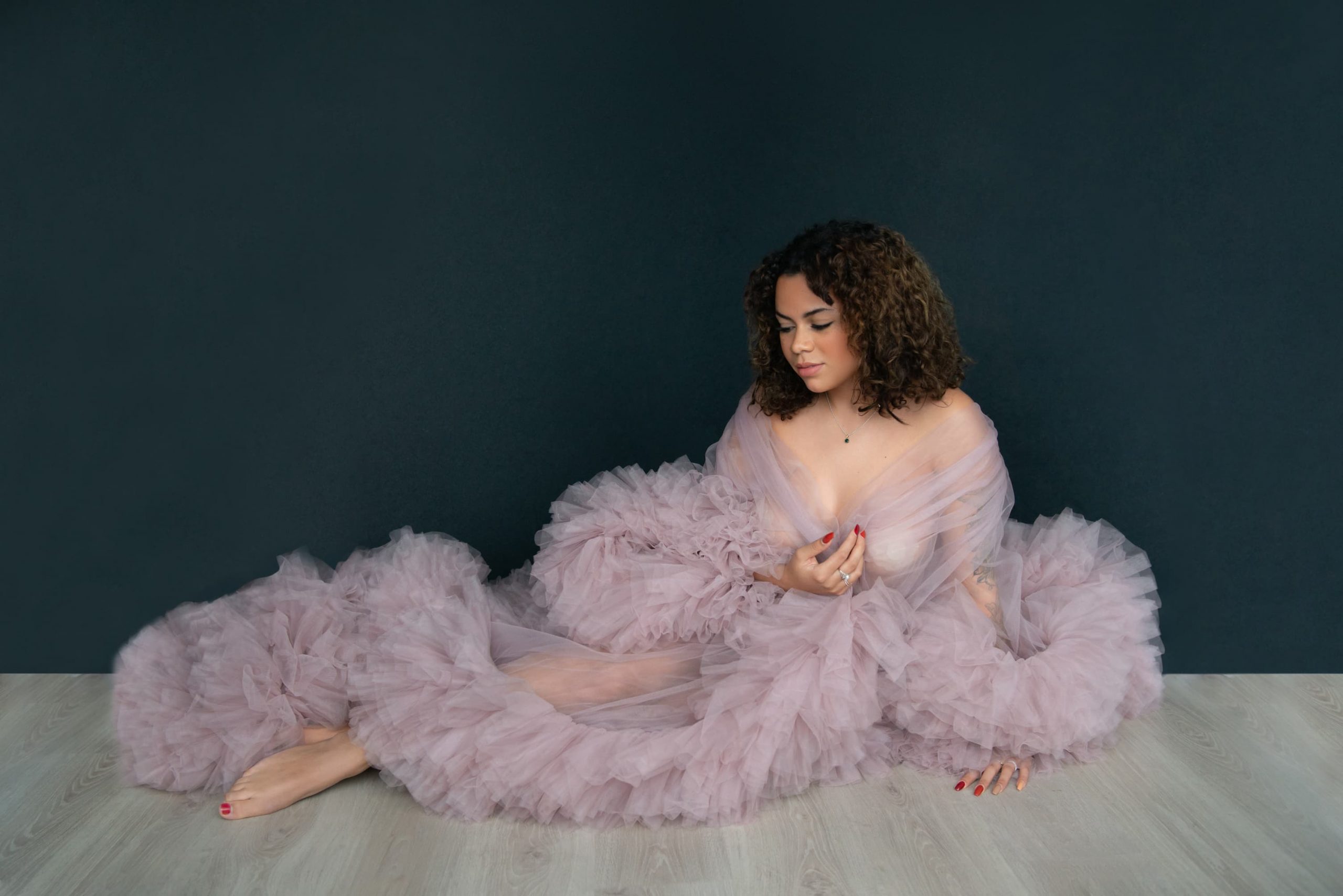 Woman with curly hair sitting on a light wooden floor against a dark wall, wrapped in a voluminous sheer pink tulle fabric, barefoot with painted toenails and fingernails, looking downward with a relaxed expression.