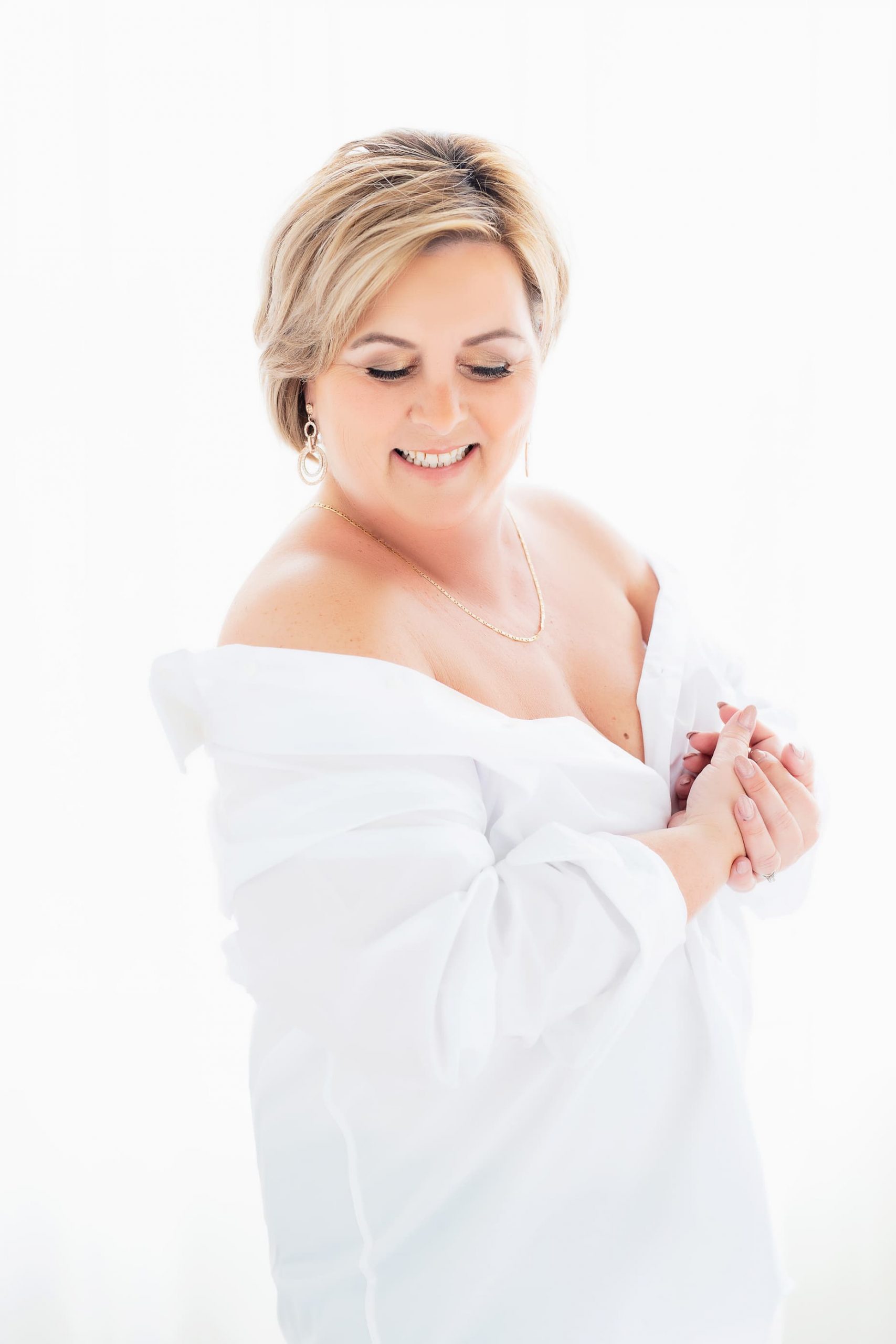 Smiling woman with short blonde hair wearing a white off-shoulder blouse, gold earrings, and a necklace, posing against a bright white background with her hands clasped.