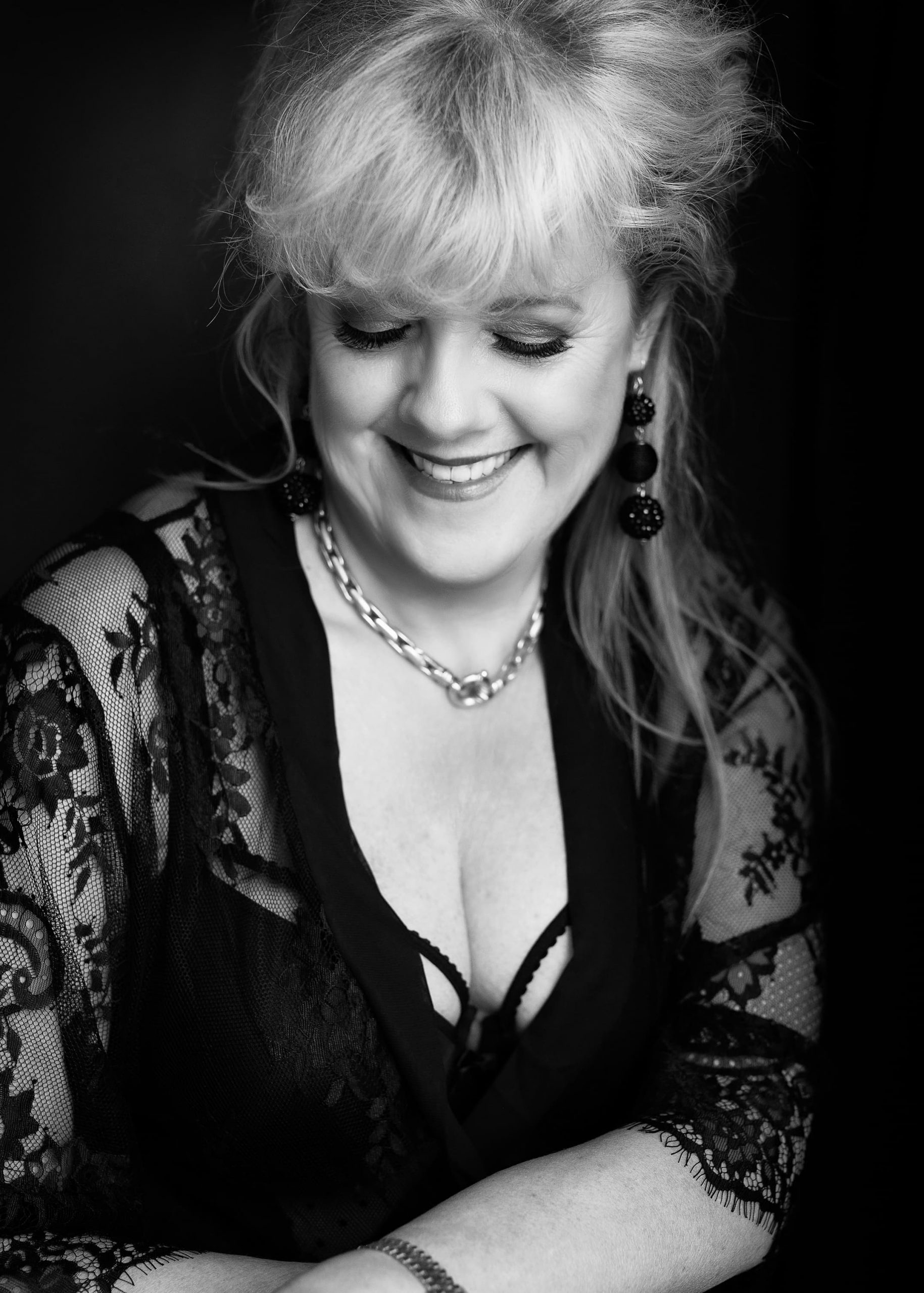 Smiling woman with closed eyes wearing a black lace top, a chunky chain necklace, elaborate earrings, and a bracelet, photographed in black and white on a dark background.