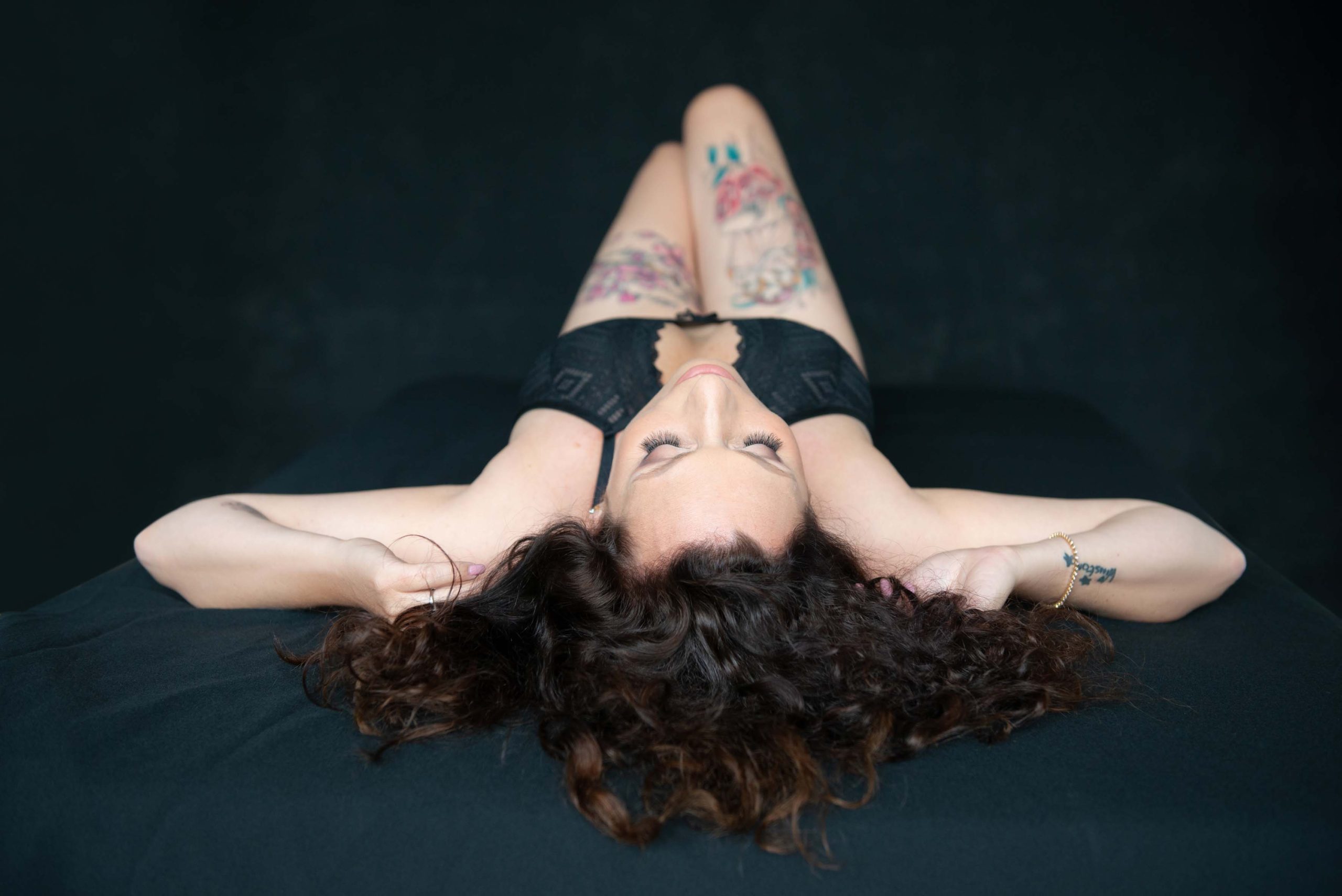 Top view of a woman lying on her back on a dark surface, wearing black lace lingerie, with long curly hair spread out and visible colorful tattoos on her thighs and a bracelet on her right wrist.