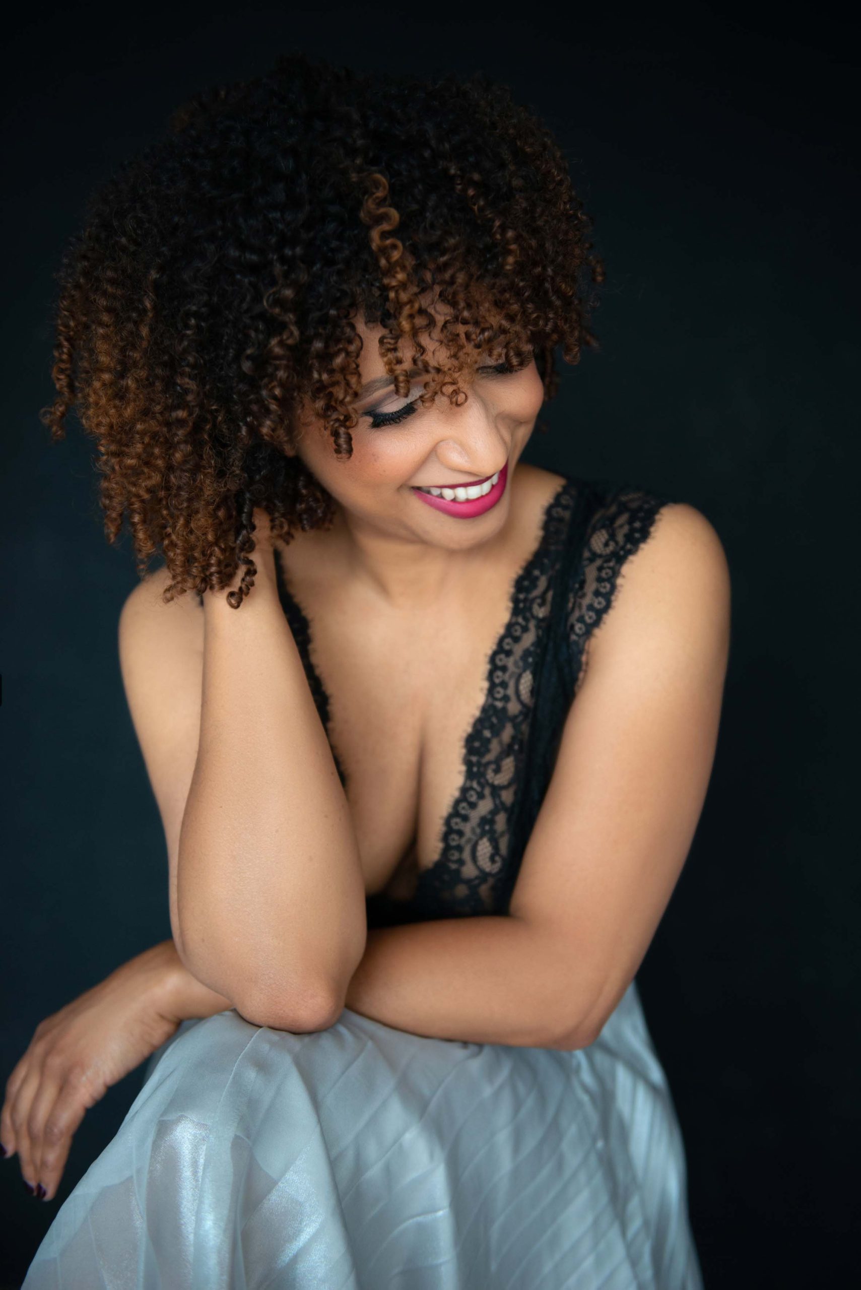 A woman with curly hair, wearing a black lace top and a light blue satin skirt, smiling and looking down with one hand touching her hair and the other resting on her knee against a dark background.