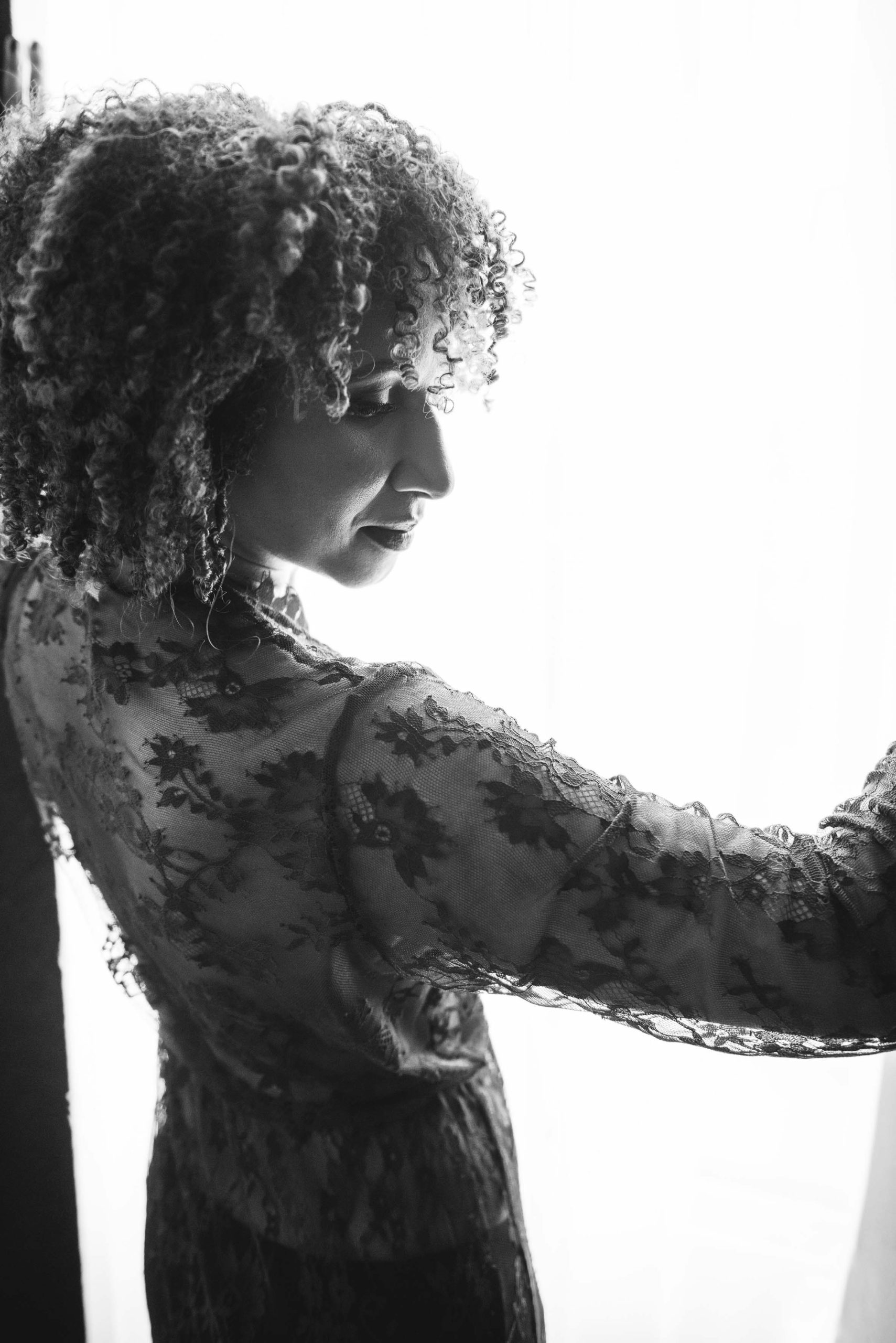 Black and white photo of a woman with curly hair wearing a sheer floral lace top, looking downward and standing near a bright window.