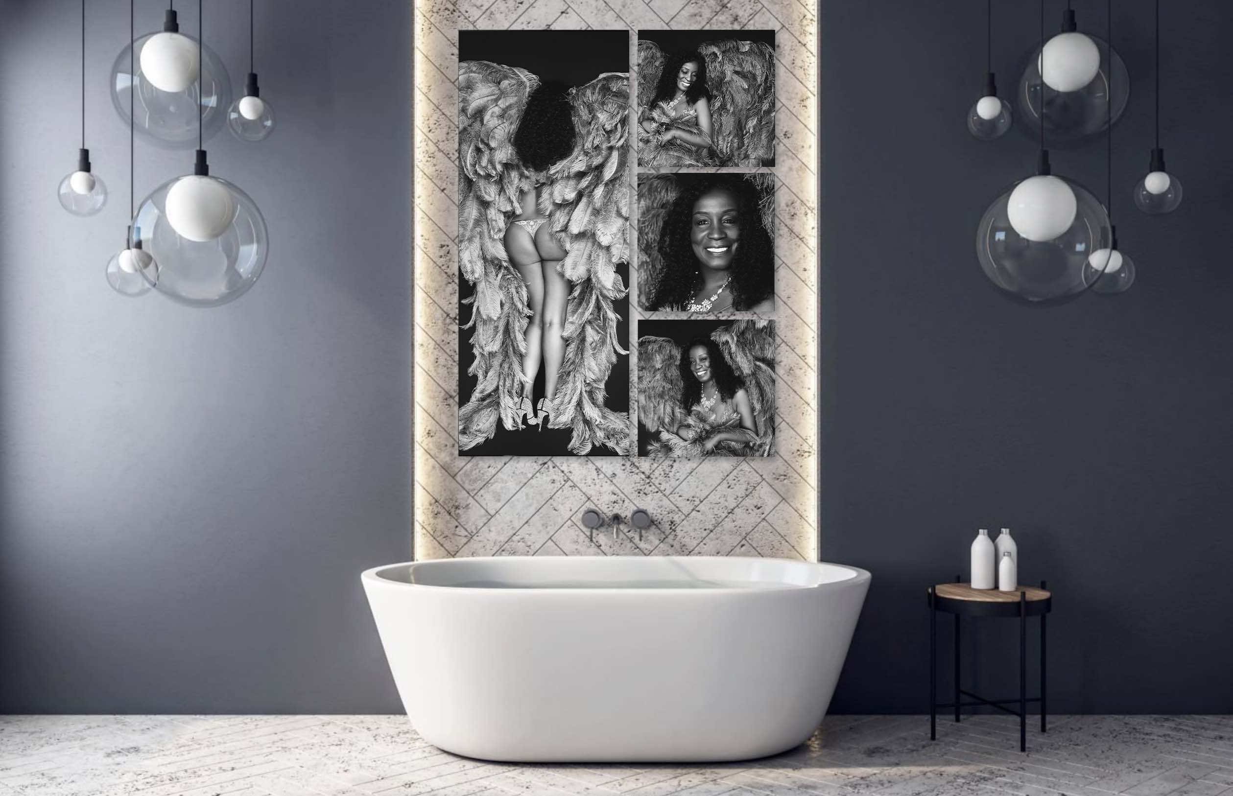 Modern bathroom with a white freestanding bathtub centered against a wall with light marble tiles and vertical LED strip lighting. Above the bathtub, four black-and-white portraits of a smiling woman with curly hair and large feathered wings form a collage. Two hanging pendant lights with translucent globe shades are on either side, and a small wooden side table with white bottles stands to the right.
